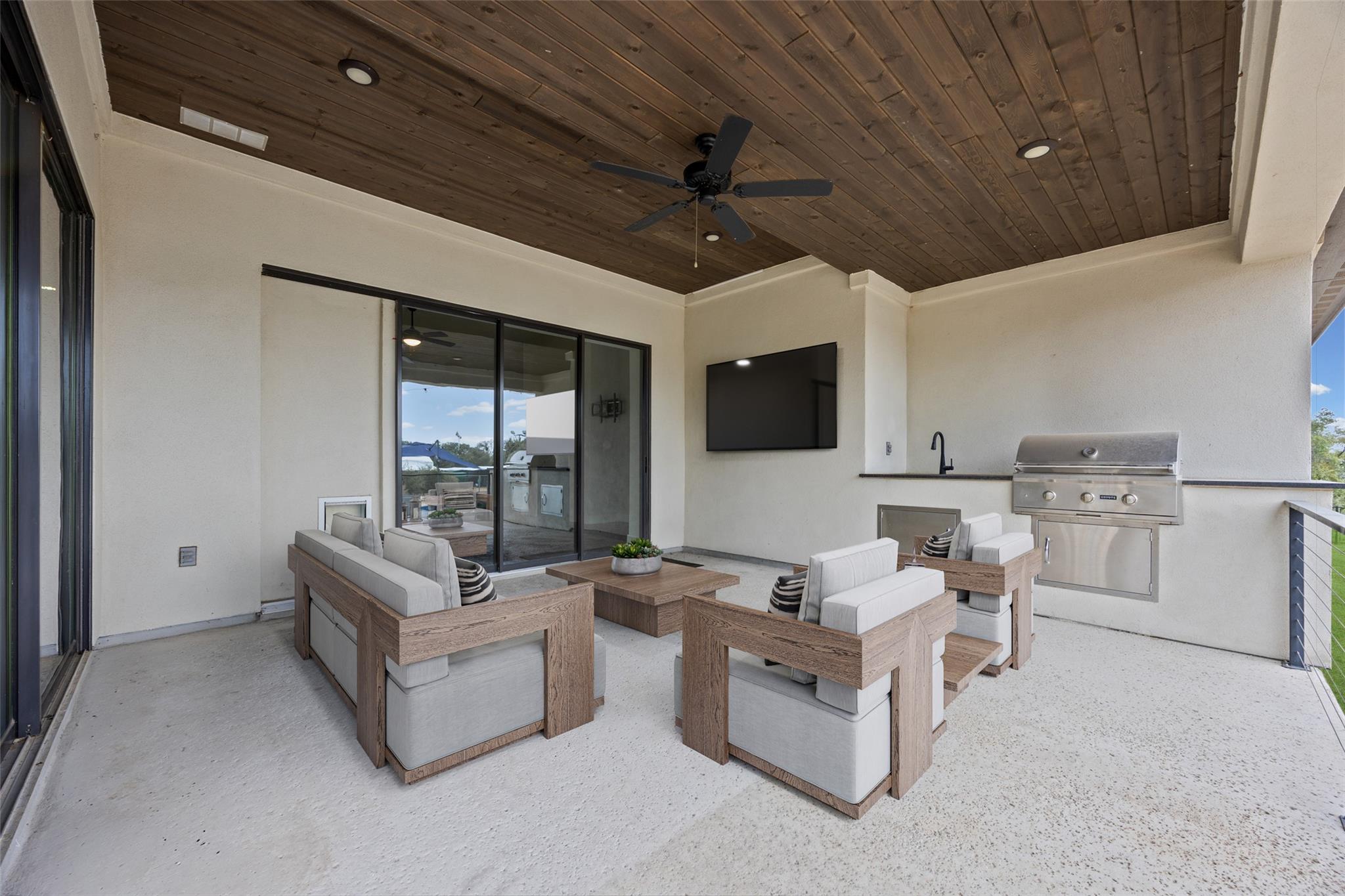 Covered patio with cushioned seating, a wood ceiling, wall-mounted TV, and built-in outdoor grill.