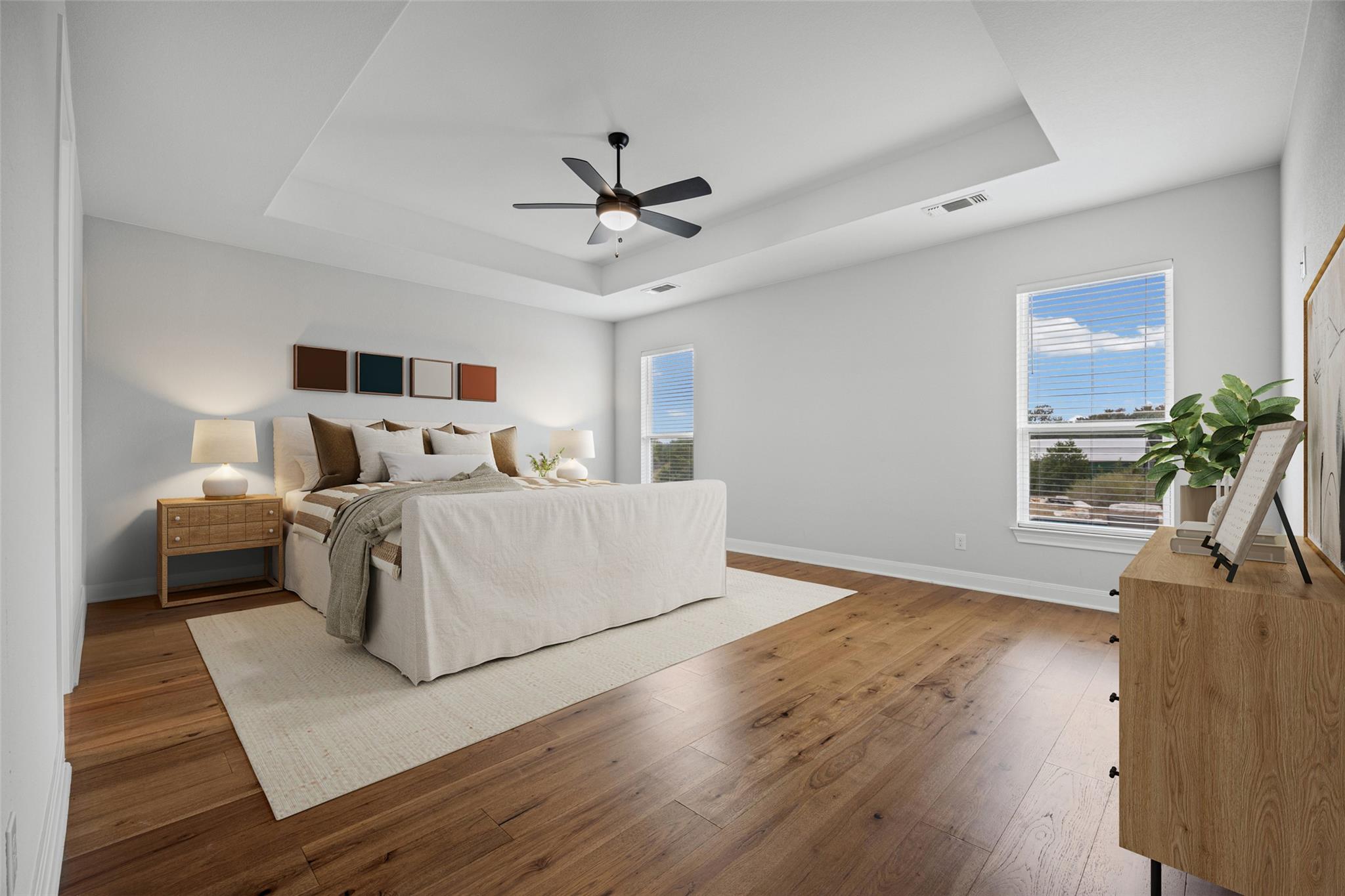 Staged bedroom with a white bed, tray ceiling, ceiling fan, and warm wood floors.