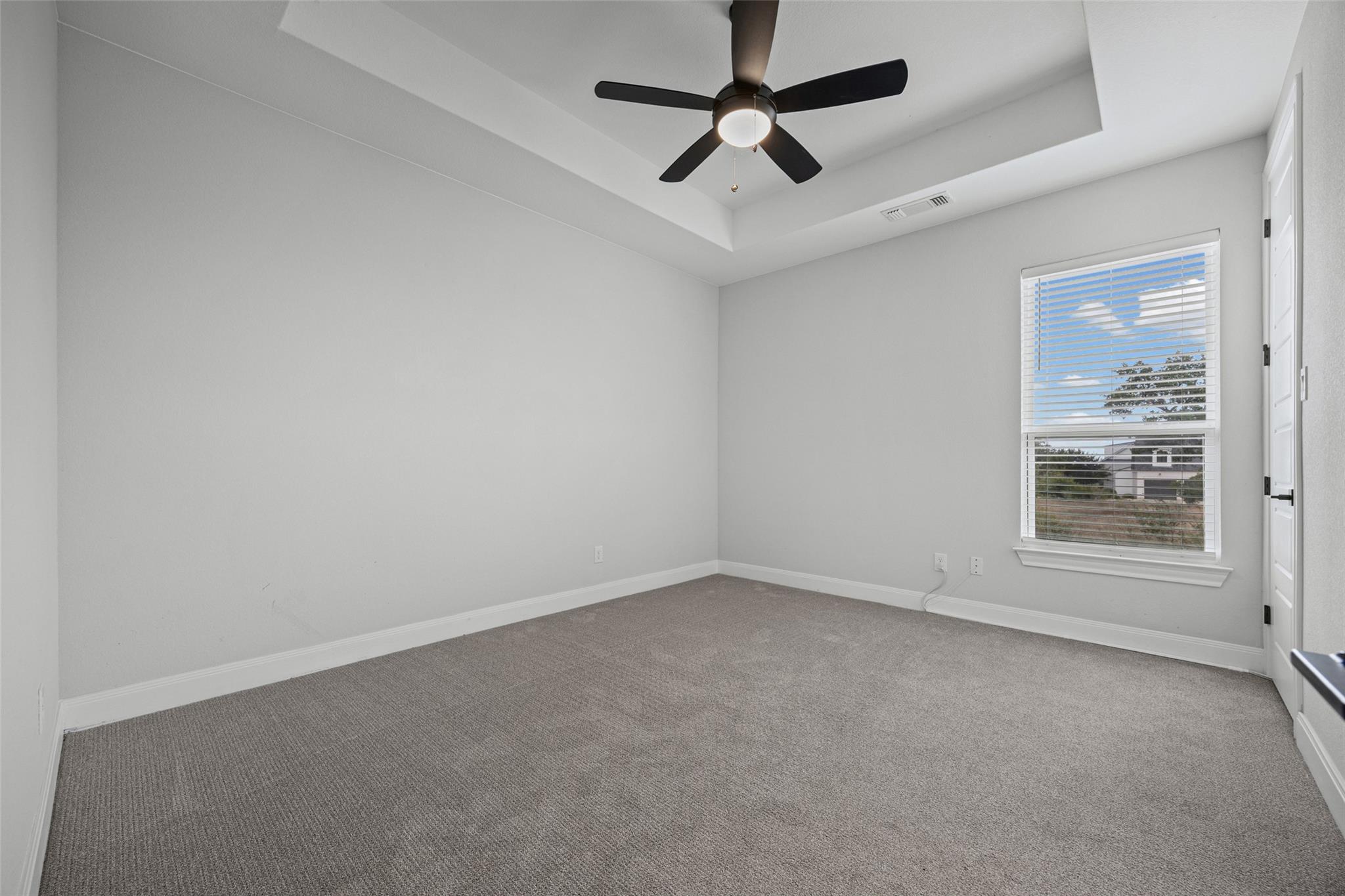 Empty bedroom with gray carpet, a tray ceiling, and a window with white blinds.