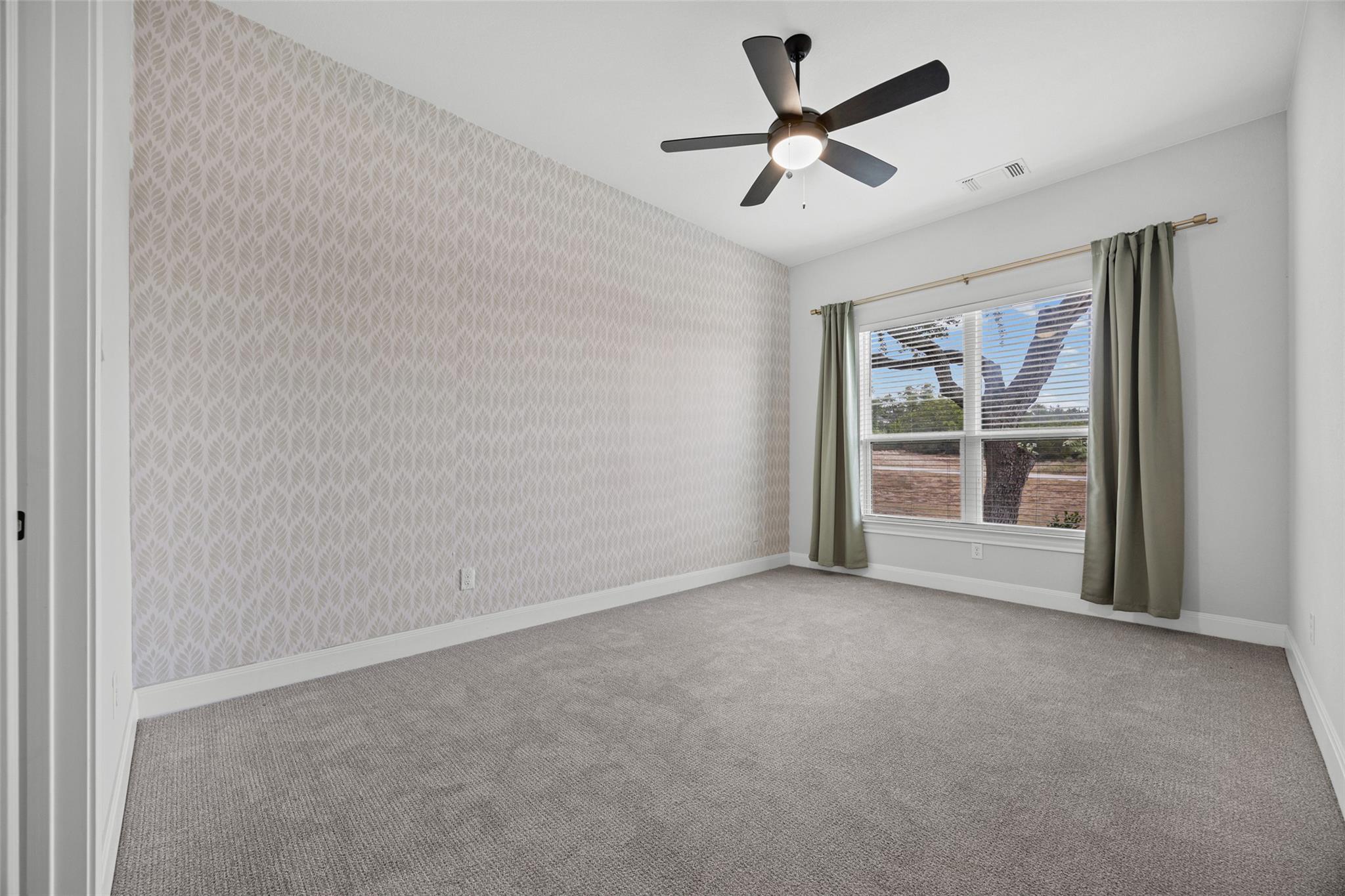 Empty bedroom with patterned wallpaper, gray carpet, and a window framed by green curtains.