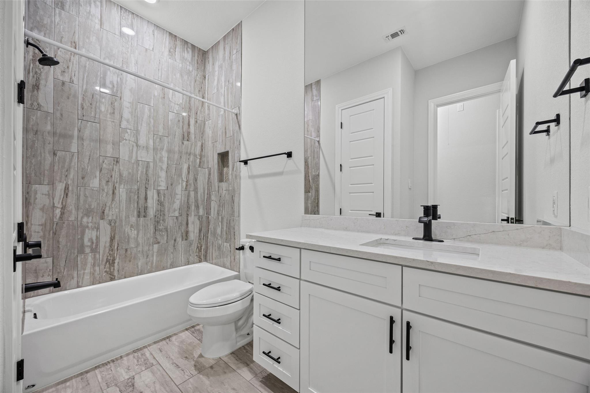 Bathroom with a gray tiled tub and shower beside a white vanity with black fixtures.