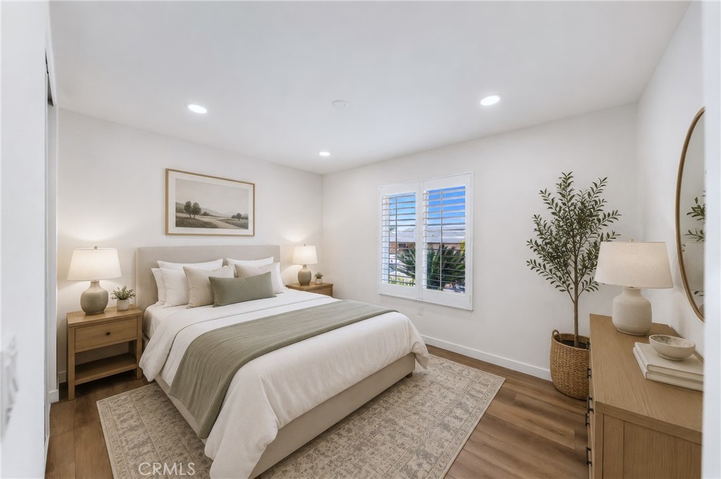 Staged bedroom with a white bed, wood nightstands, shuttered window, and neutral decor.