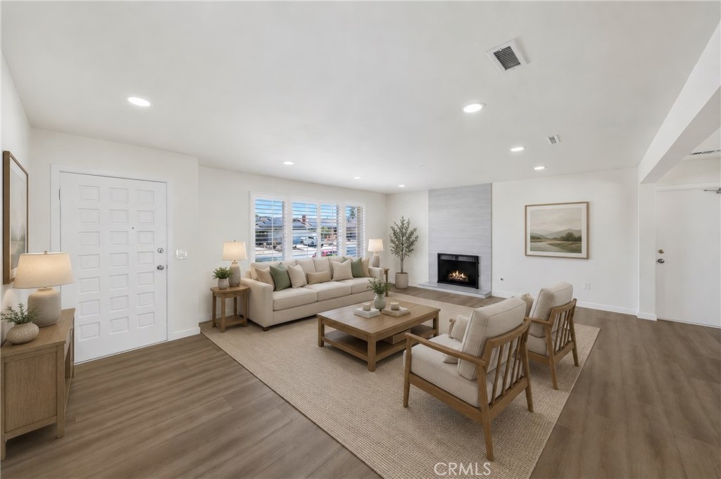Staged living room with a lit fireplace, beige seating, wide shuttered windows, and wood-look floors.