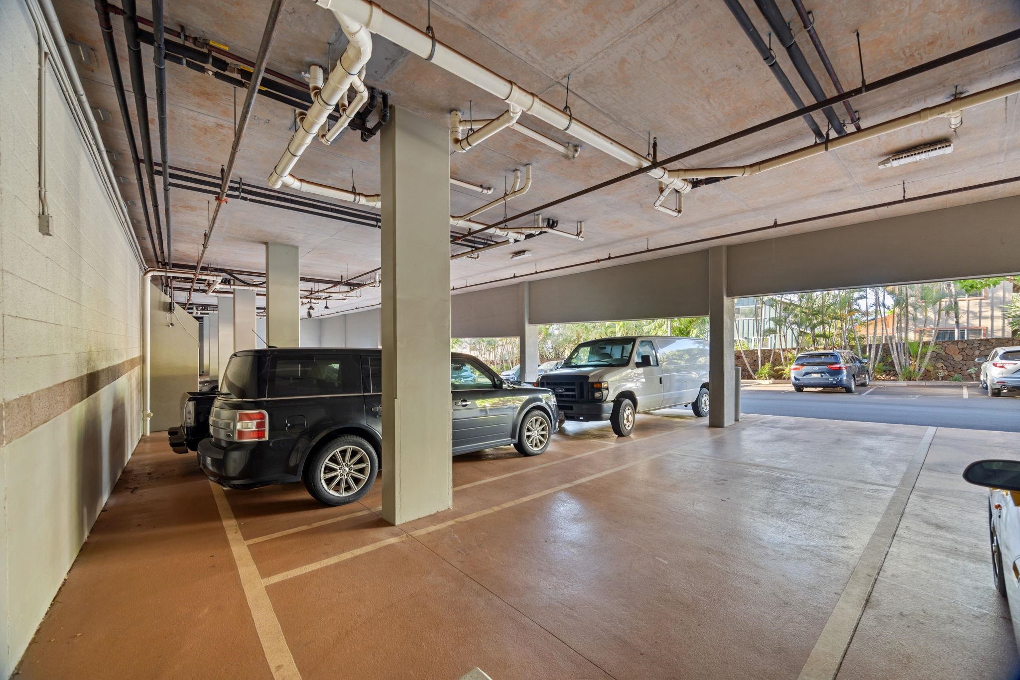 Covered parking area with marked spaces, exposed pipes overhead, and several parked vehicles.