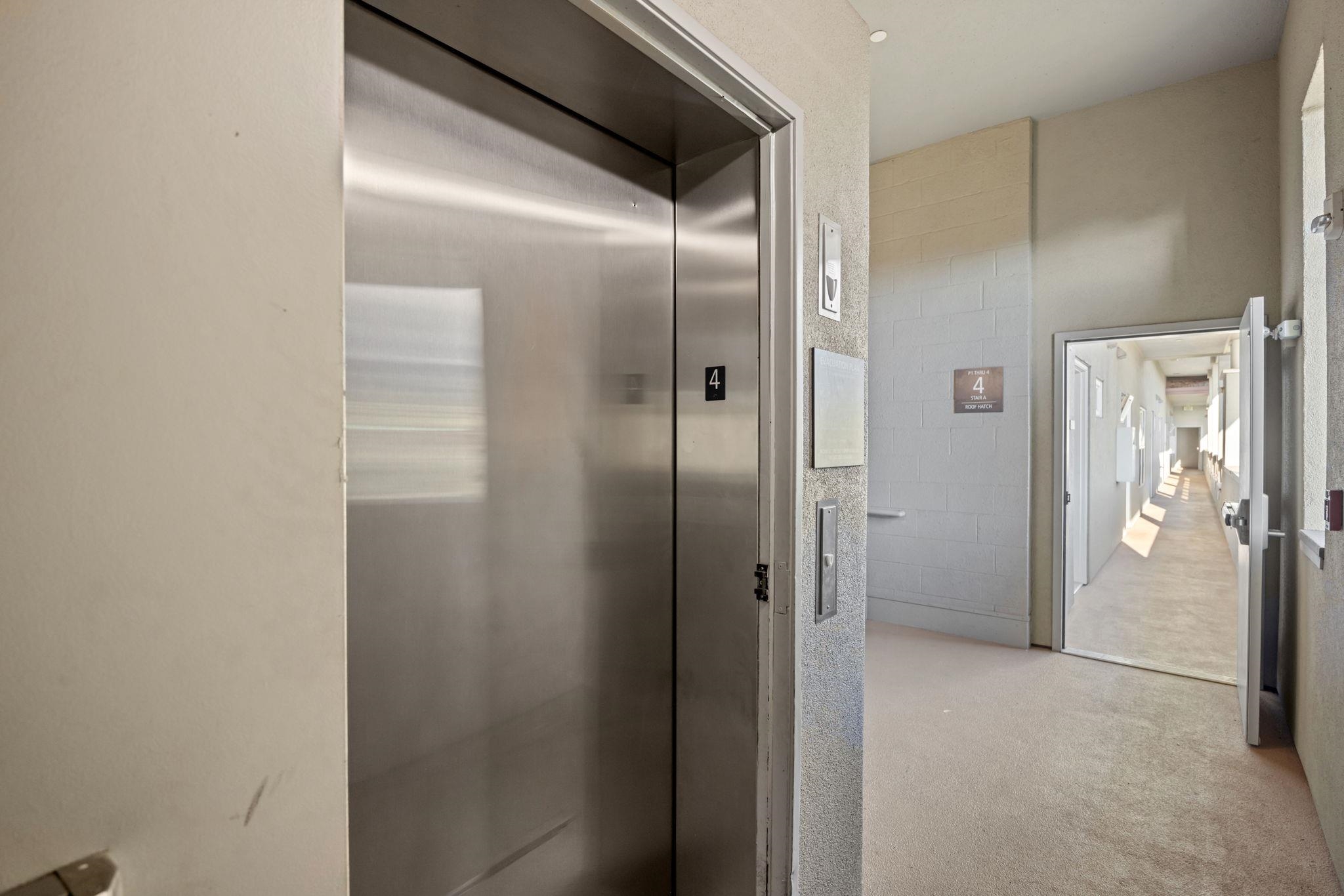 Elevator lobby with stainless-steel doors and an open doorway to a sunlit corridor.