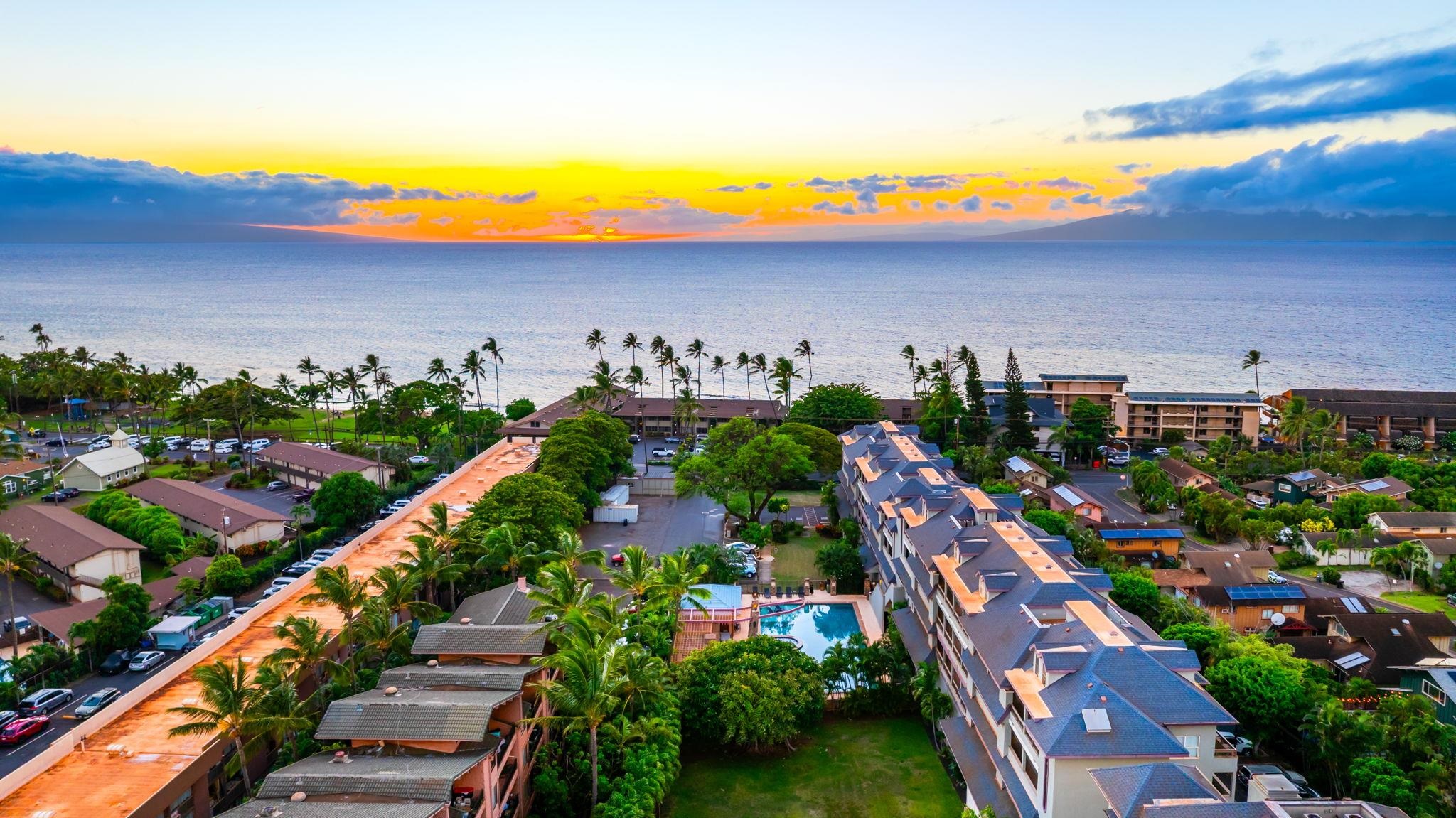Aerial view of coastal condominiums and a pool beneath a colorful sunset over the Pacific Ocean.
