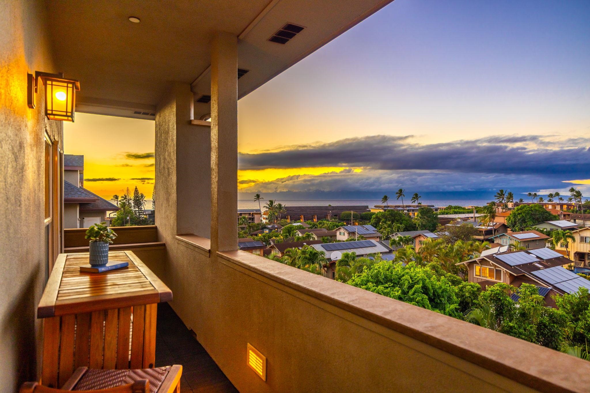 Covered balcony overlooking a palm-lined neighborhood and ocean at sunset, with a wooden table and potted plant.