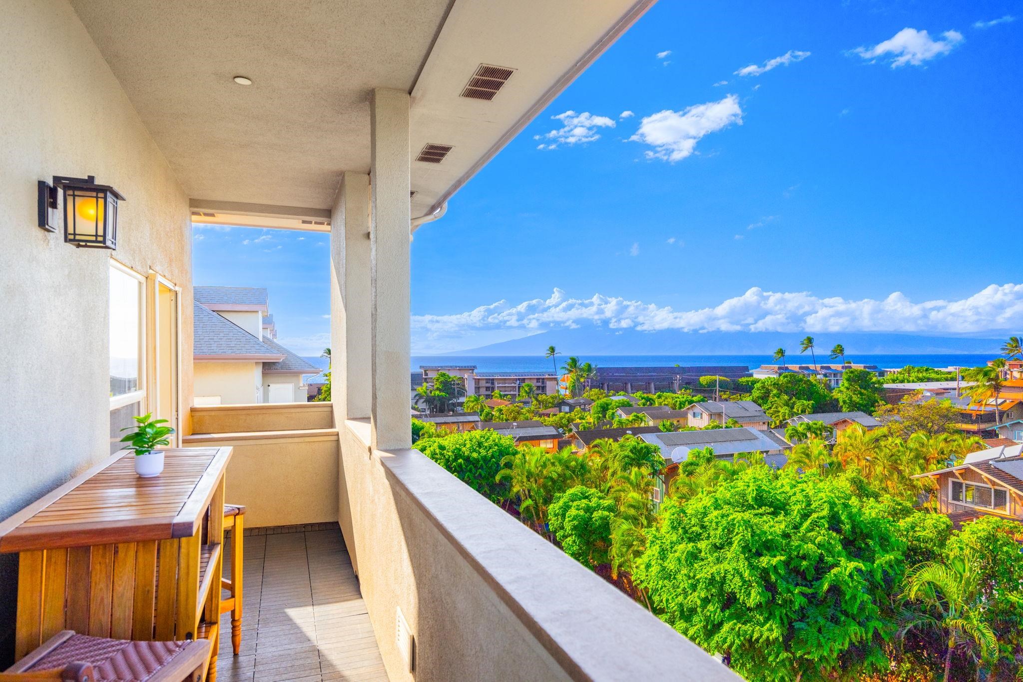 Covered balcony with a wooden table overlooks palm-covered homes, ocean, and a distant island.