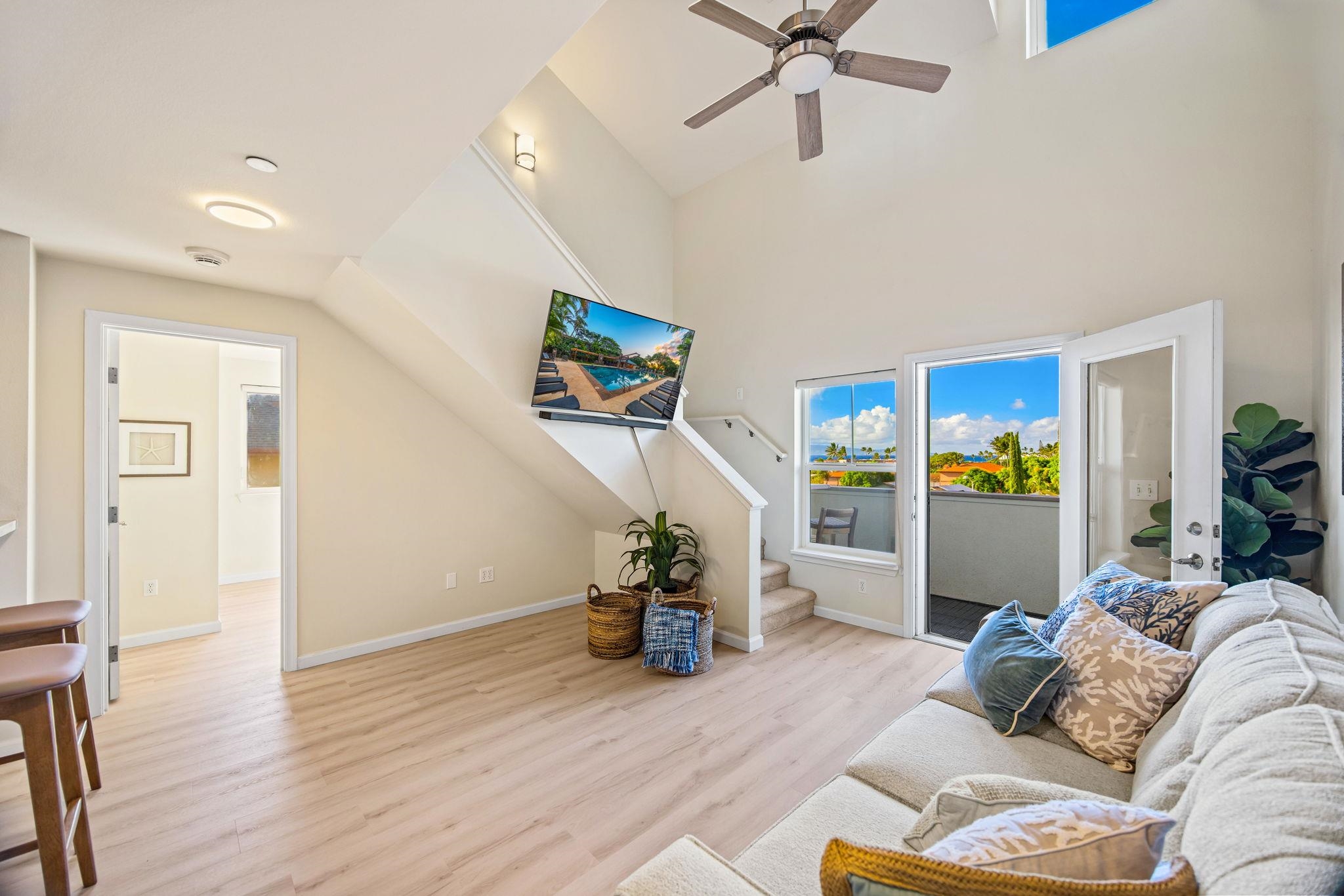 Bright living room with vaulted ceiling, sectional sofa, and open balcony door overlooking tropical greenery.