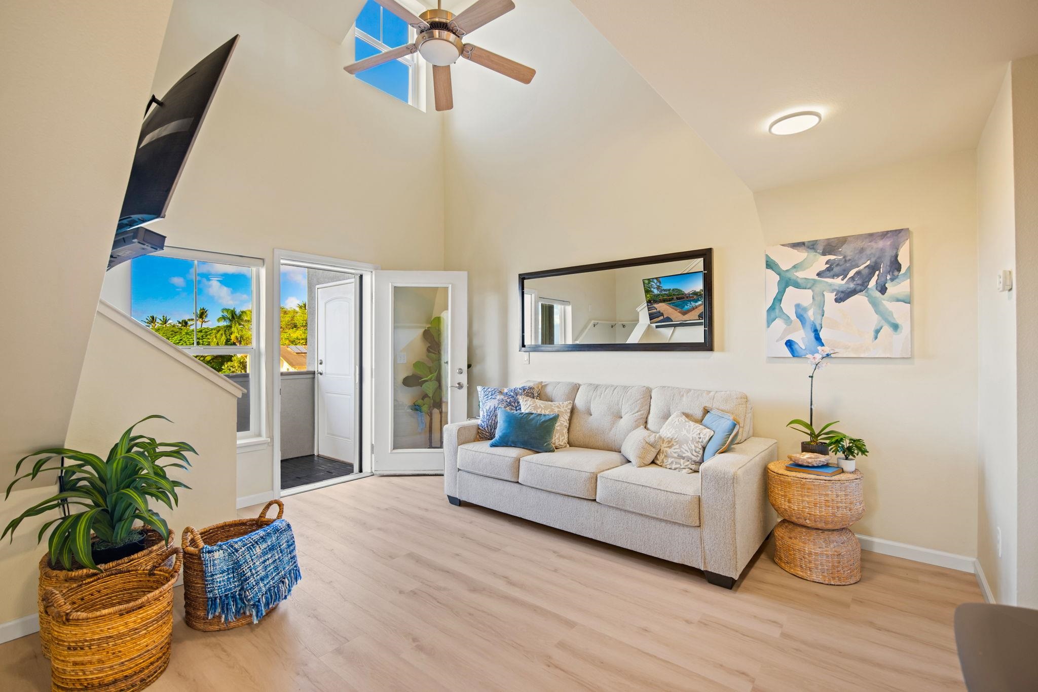 Living room with a vaulted ceiling, ceiling fan, and balcony doors overlooking tropical greenery.