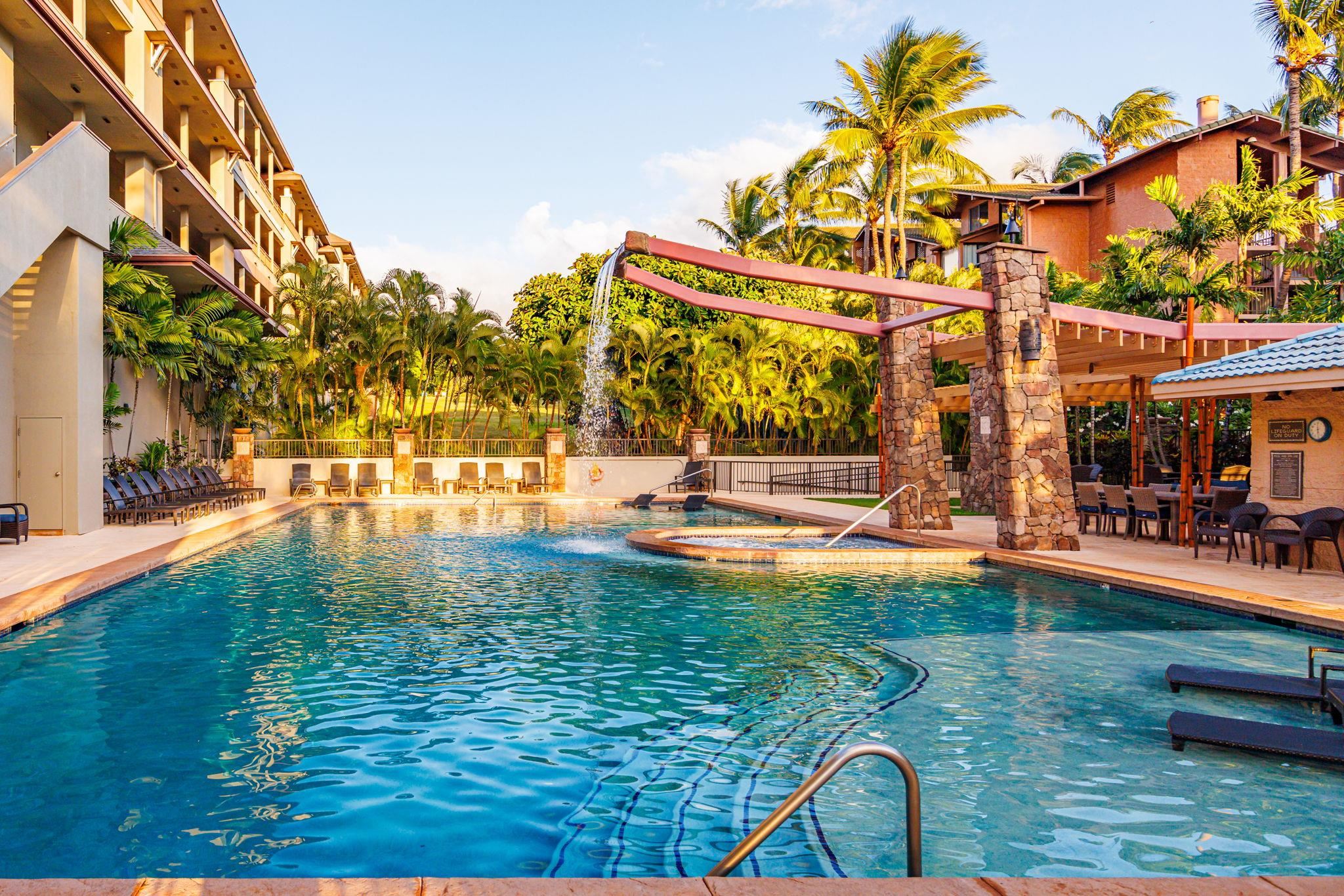 Resort pool with a stone water feature, palm trees, and lounge chairs between buildings.