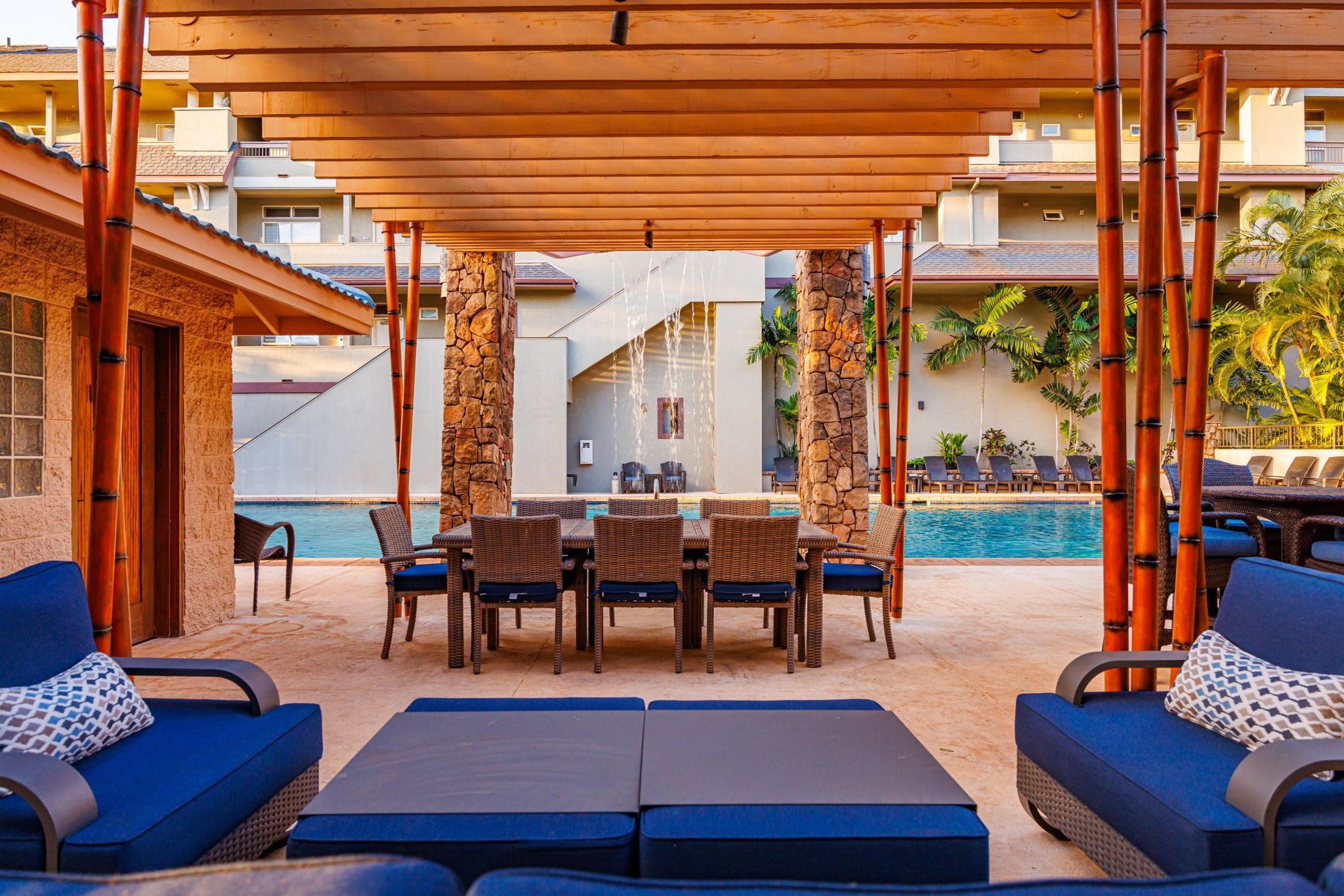 Resort pool patio with a pergola-covered dining table, blue lounge seating, fountain, and palm trees.