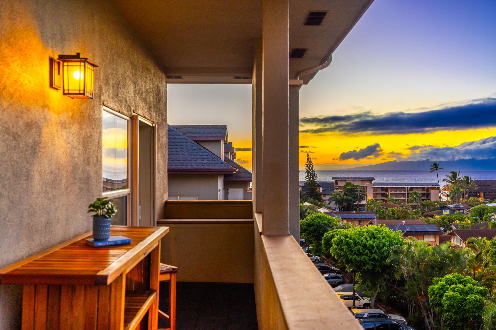 Furnished balcony overlooking palm-lined homes and the ocean beneath a vivid sunset.