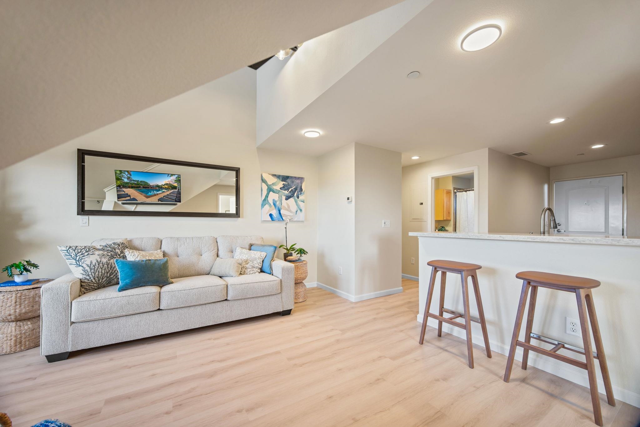 Furnished condo living room with a light sofa, blue accents, and a kitchen island with two stools.
