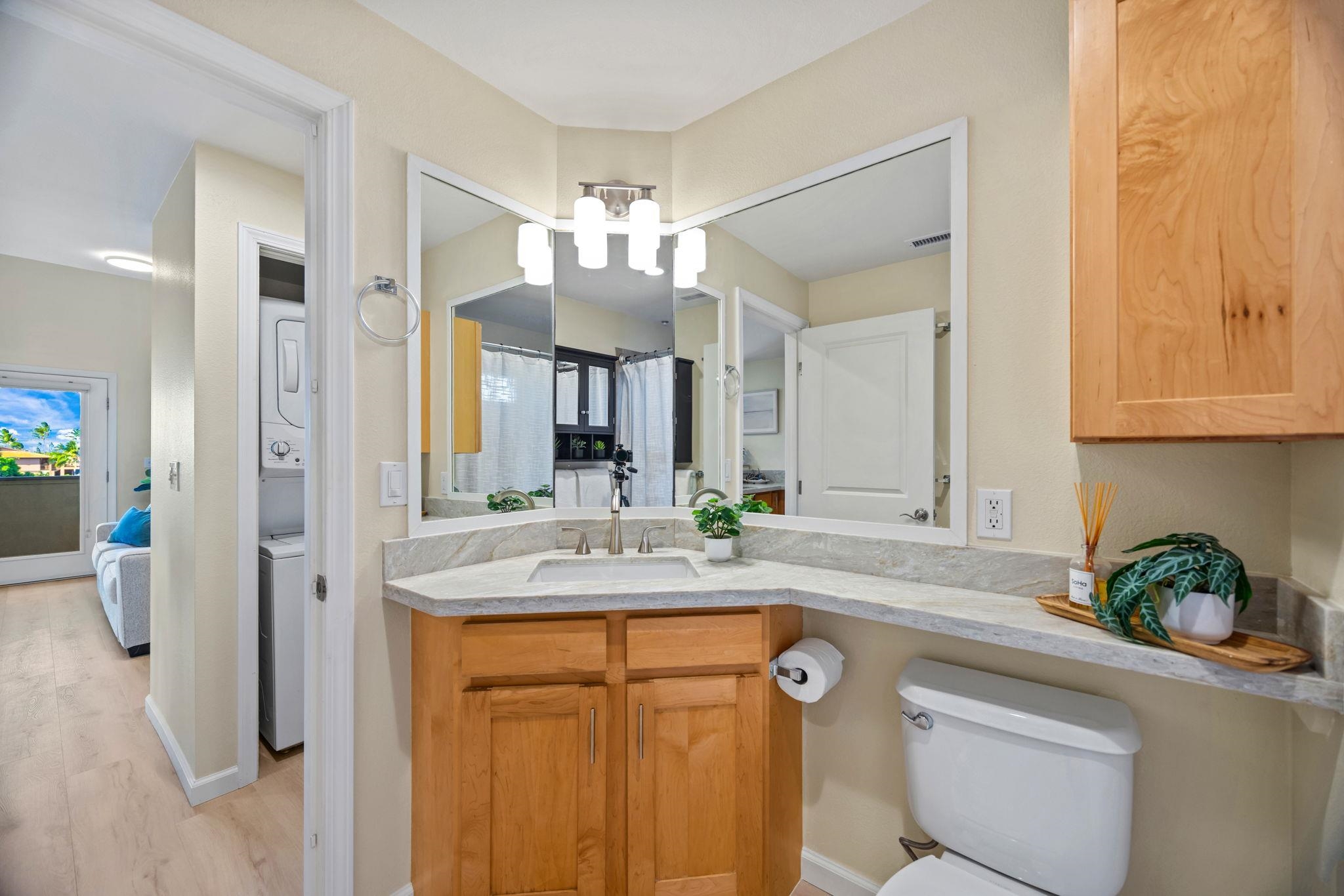 Bathroom with a stone vanity, large mirrors, light wood cabinetry, and a washer and dryer visible through the doorway.