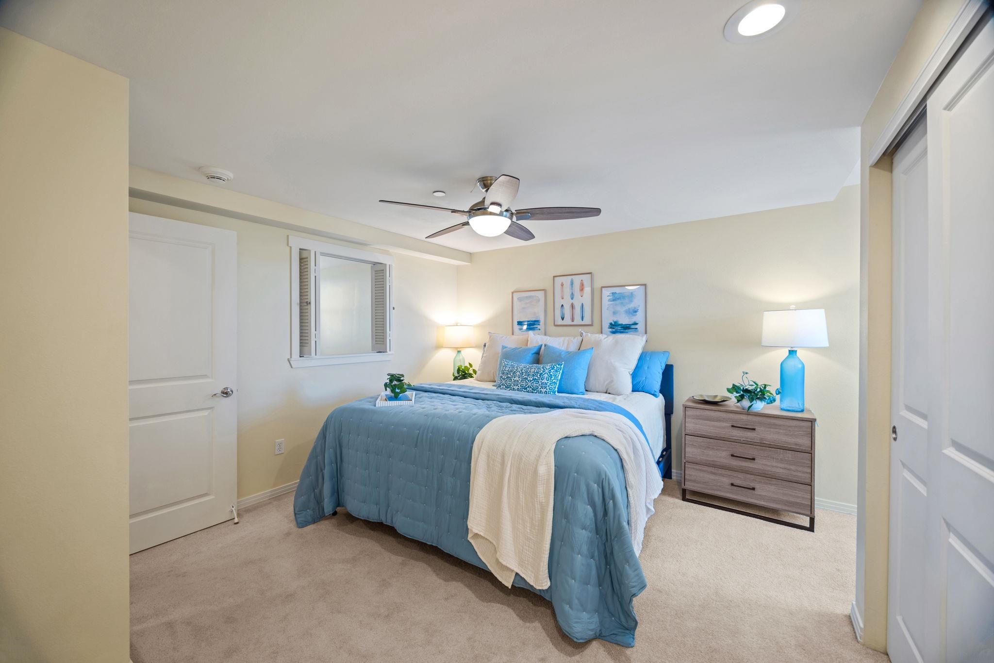 Staged bedroom with a blue-covered bed, coastal artwork, ceiling fan, and wooden dresser.