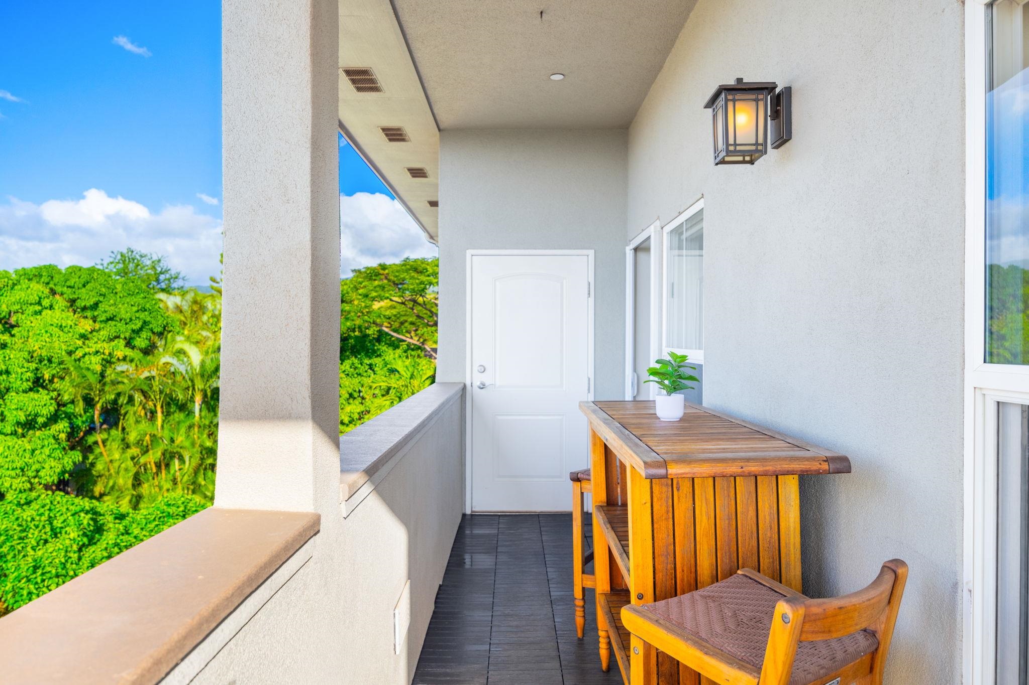 Covered balcony furnished with a wooden table and chair, overlooking lush tropical trees.