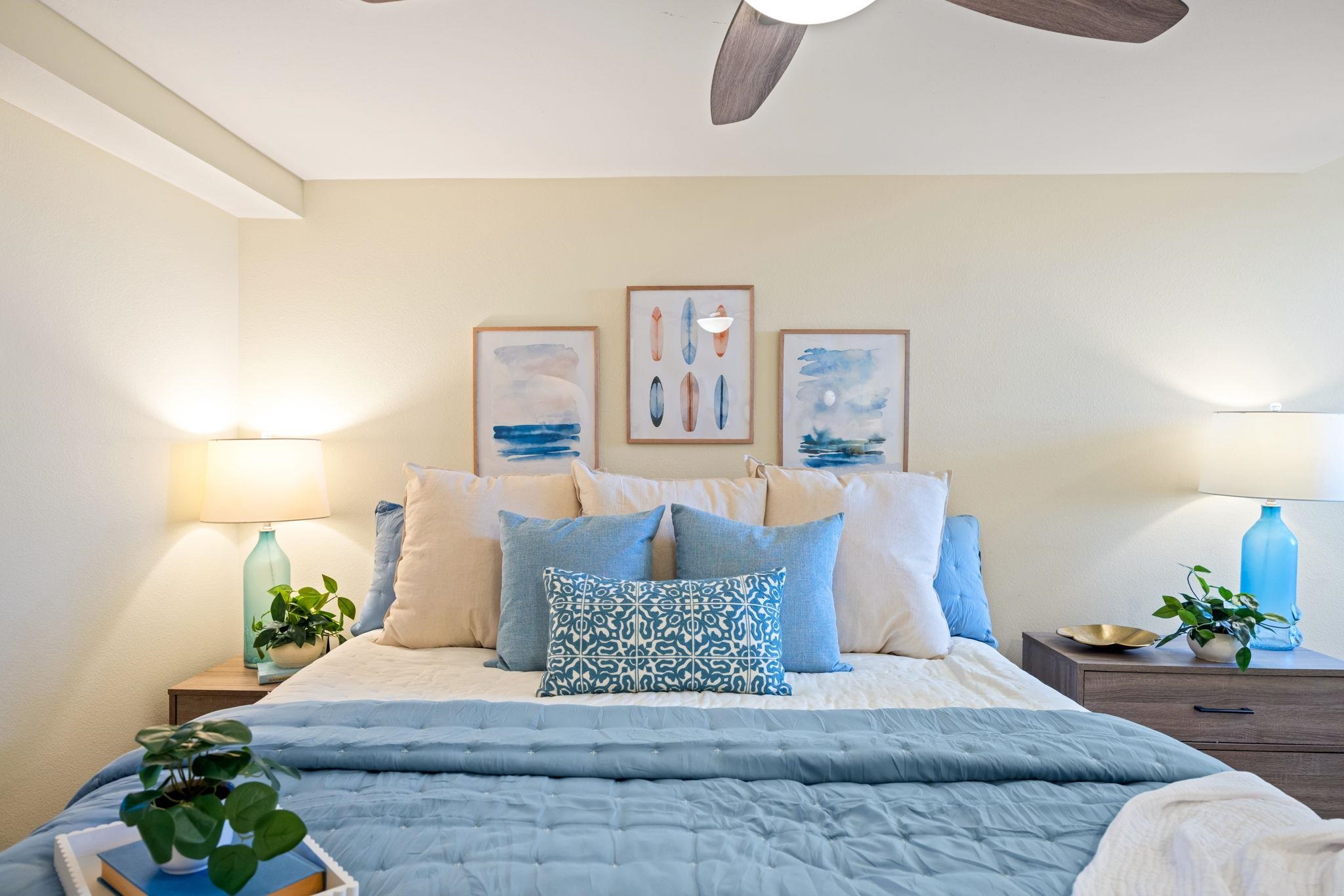 Staged bedroom with a pale blue quilt, layered pillows, bedside lamps, and coastal artwork.