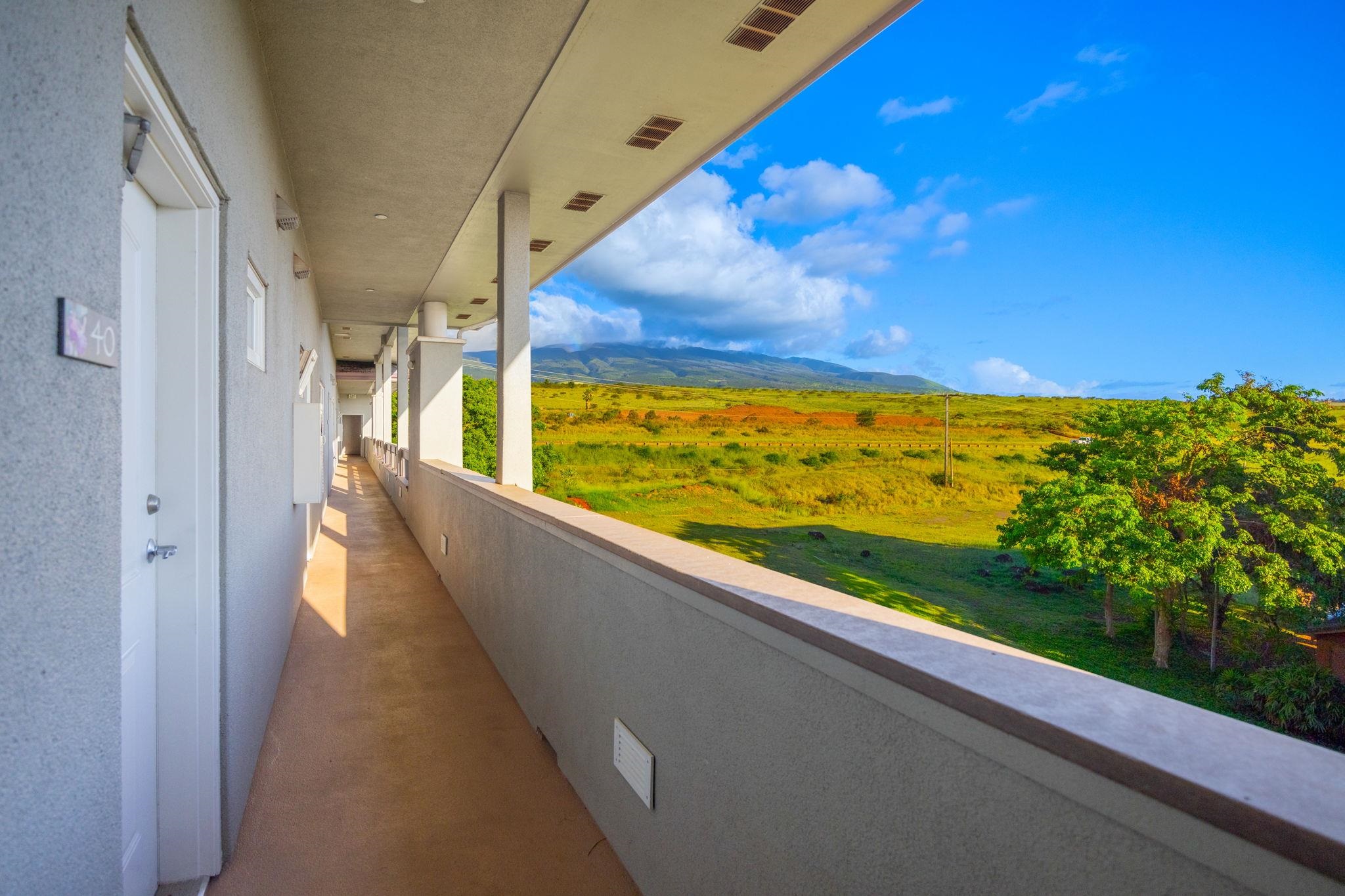 Covered apartment corridor overlooks green grounds and distant mountains beneath a bright blue sky.
