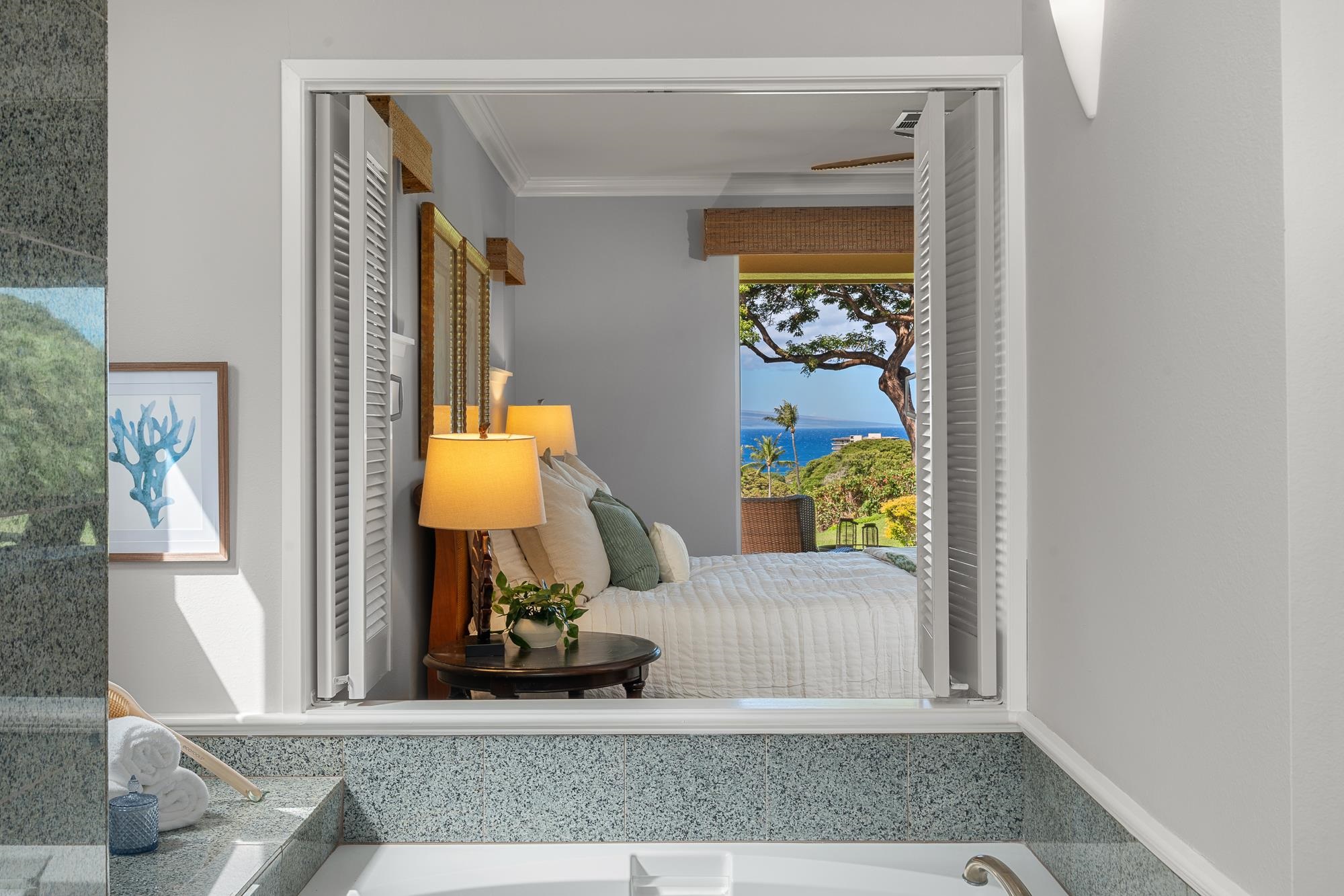 Tiled soaking tub with a wide window framing a bedroom, tropical greenery, and ocean view.