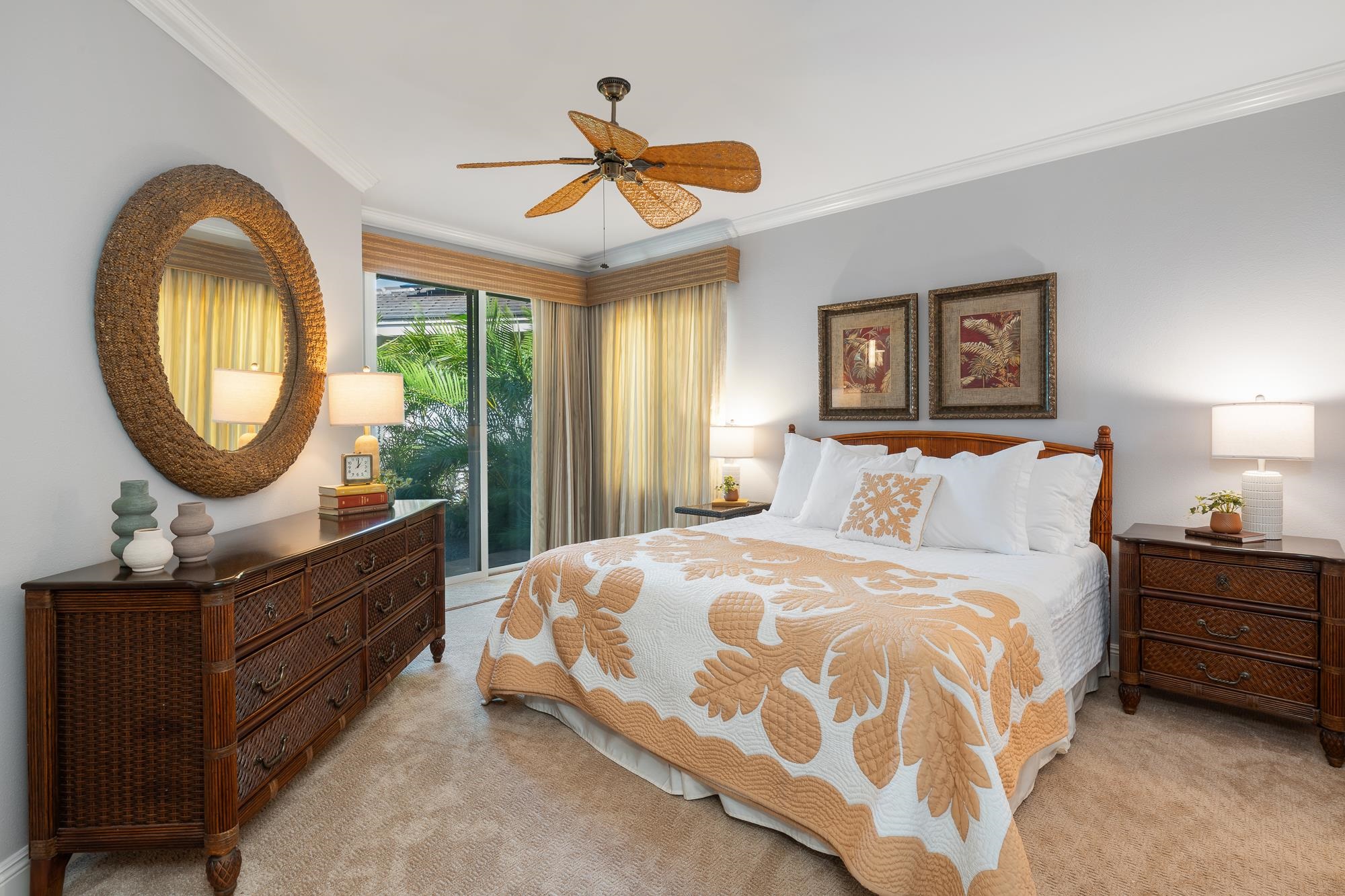 Furnished bedroom with a cream-and-tan floral quilt, woven wood furniture, and sliding doors overlooking tropical greenery.