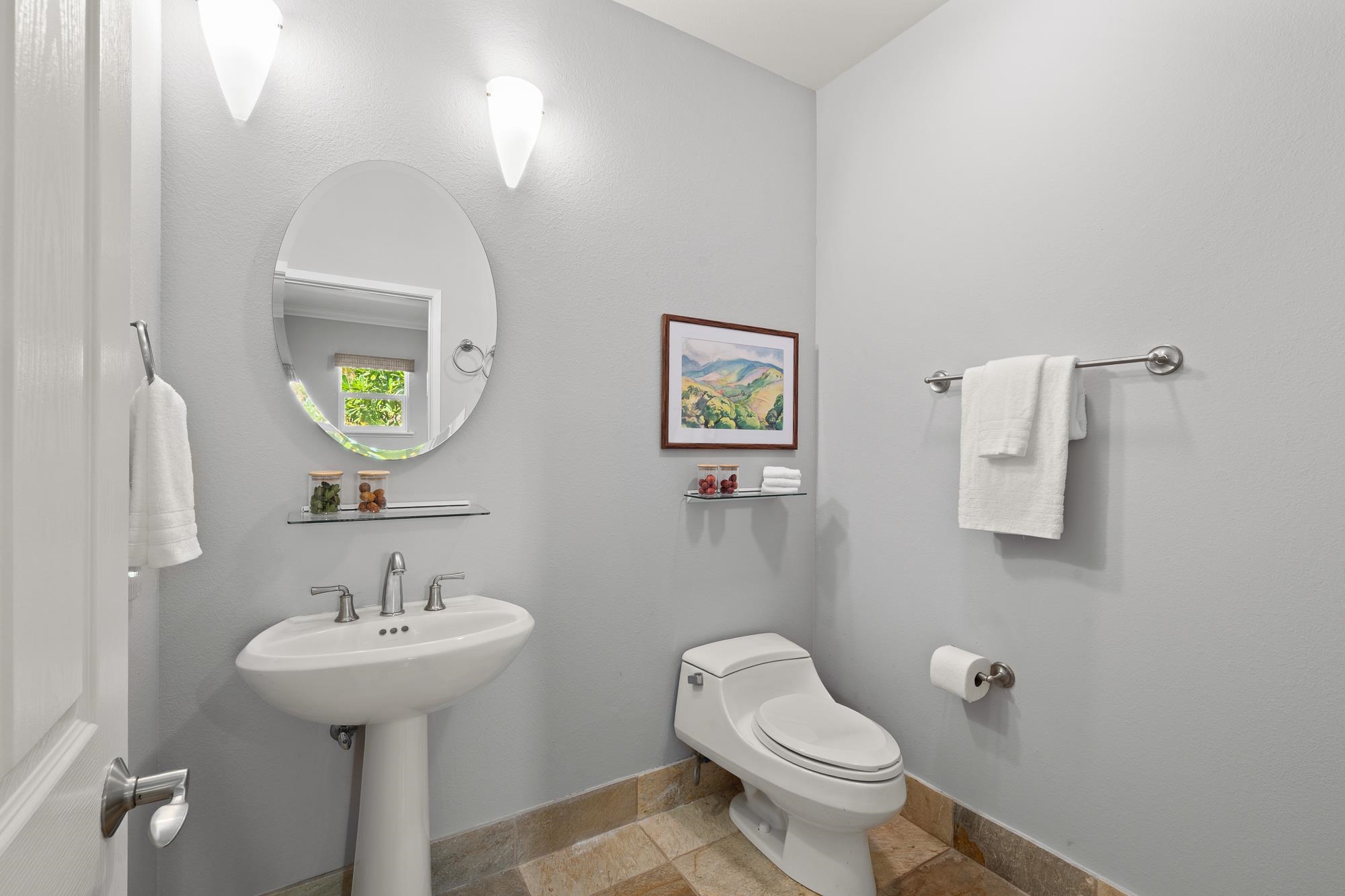 Bathroom with a pedestal sink, oval mirror, toilet, and tan stone tile flooring.