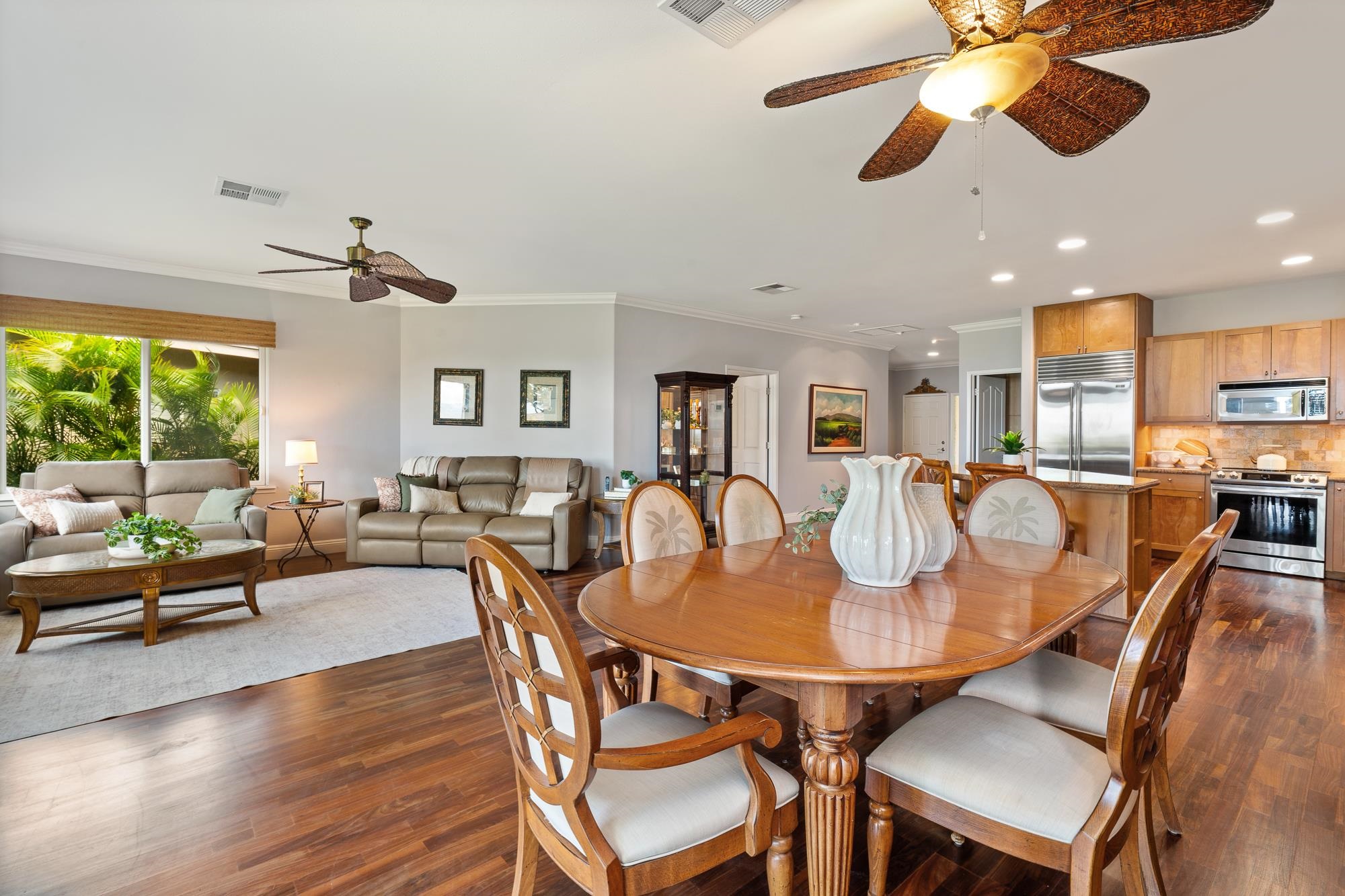 Open-plan living and dining room with a large wooden table and a window overlooking tropical greenery.