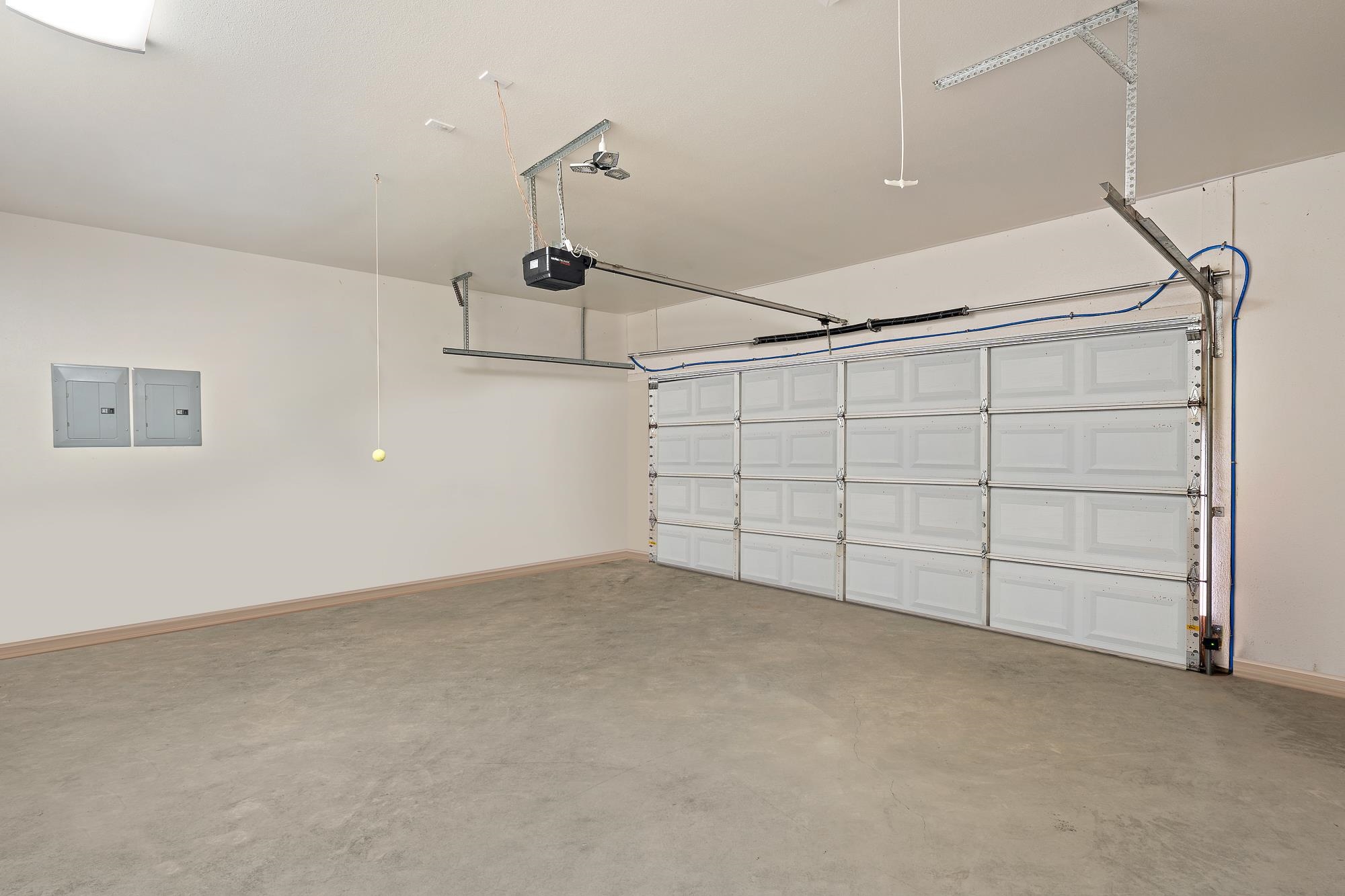 Empty garage with a wide sectional door, overhead opener, electrical panels, and smooth concrete floor.