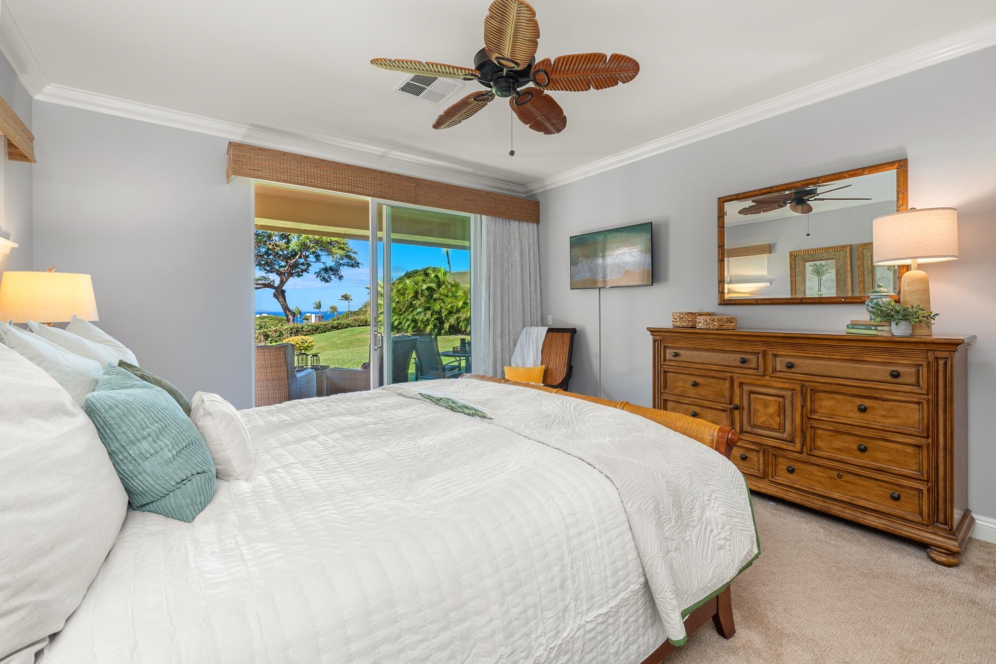 Furnished bedroom with a white bed and wooden dresser, opening onto a garden with an ocean glimpse.