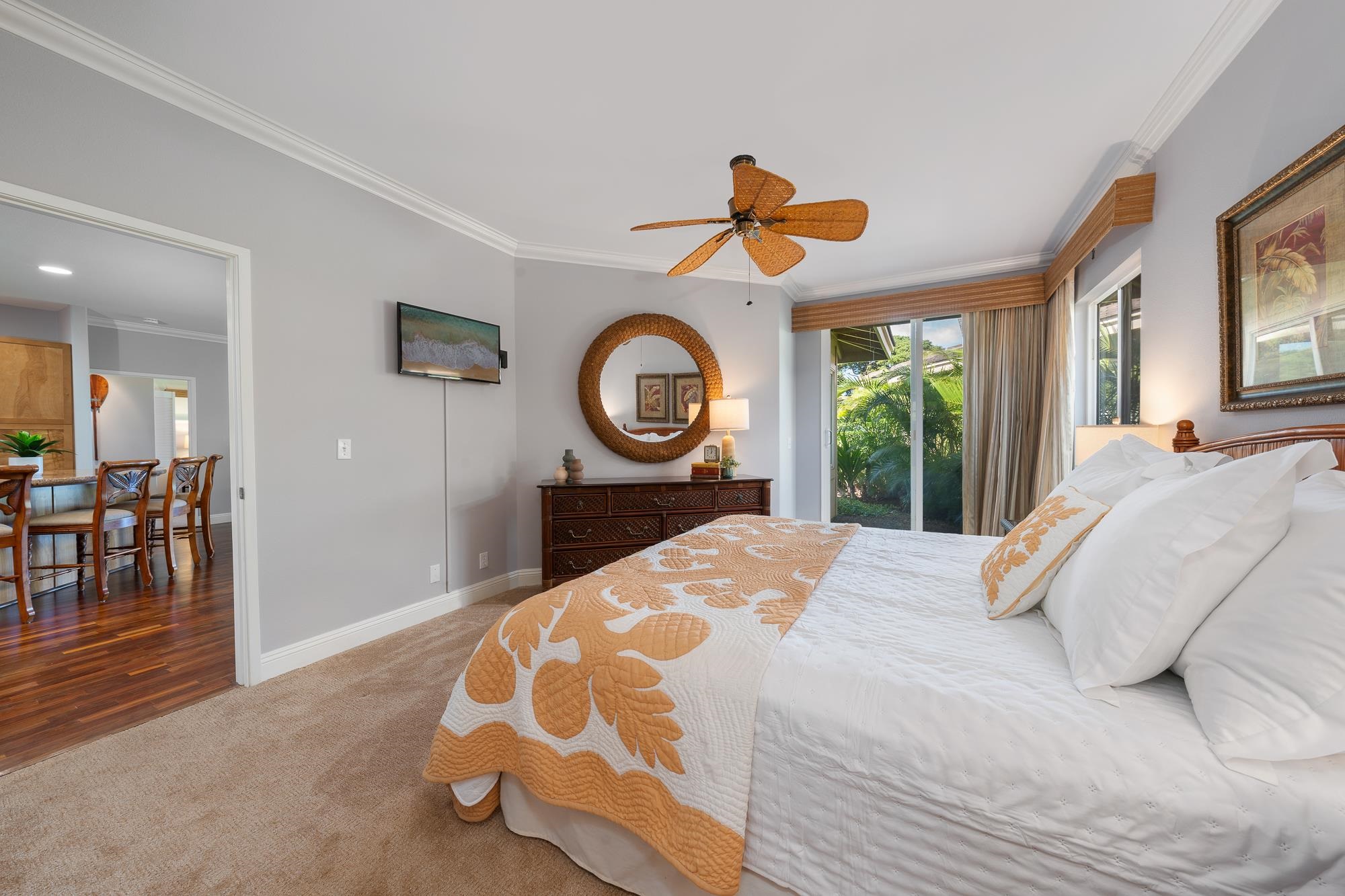 Bedroom with a gold-patterned quilt, woven ceiling fan, and sliding glass doors overlooking tropical greenery.