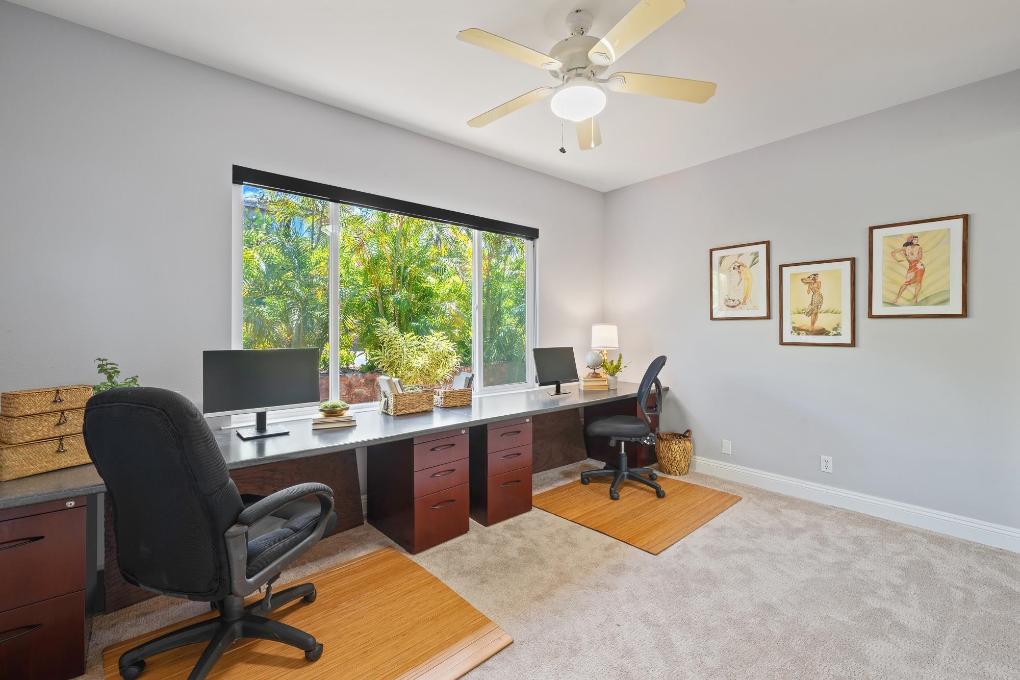 Furnished home office with a long desk and large window overlooking tropical greenery.