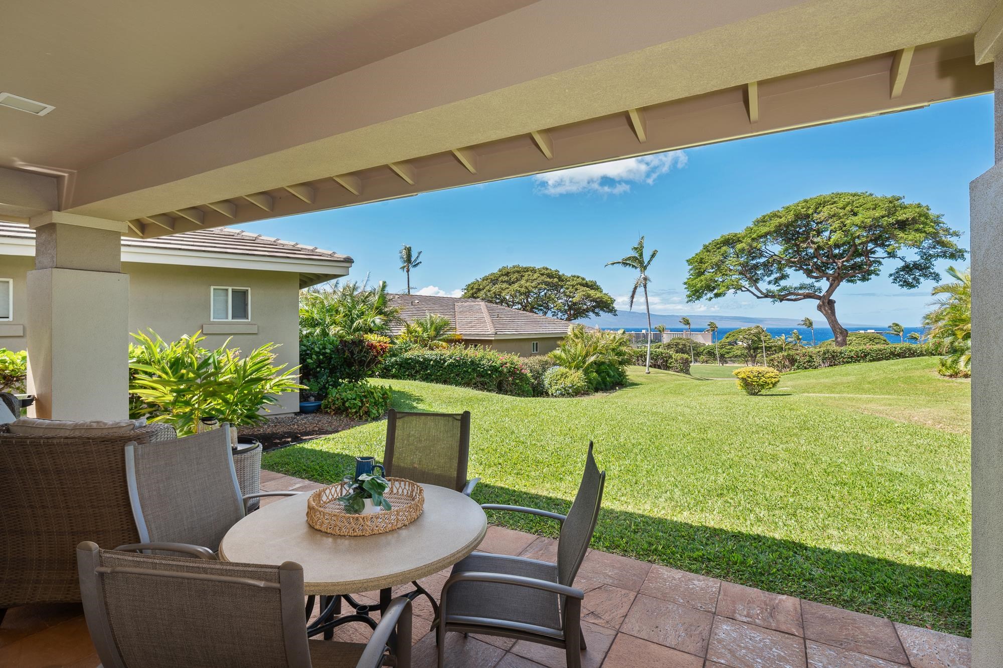 Covered patio dining area overlooking a green lawn, tropical trees, and the ocean.