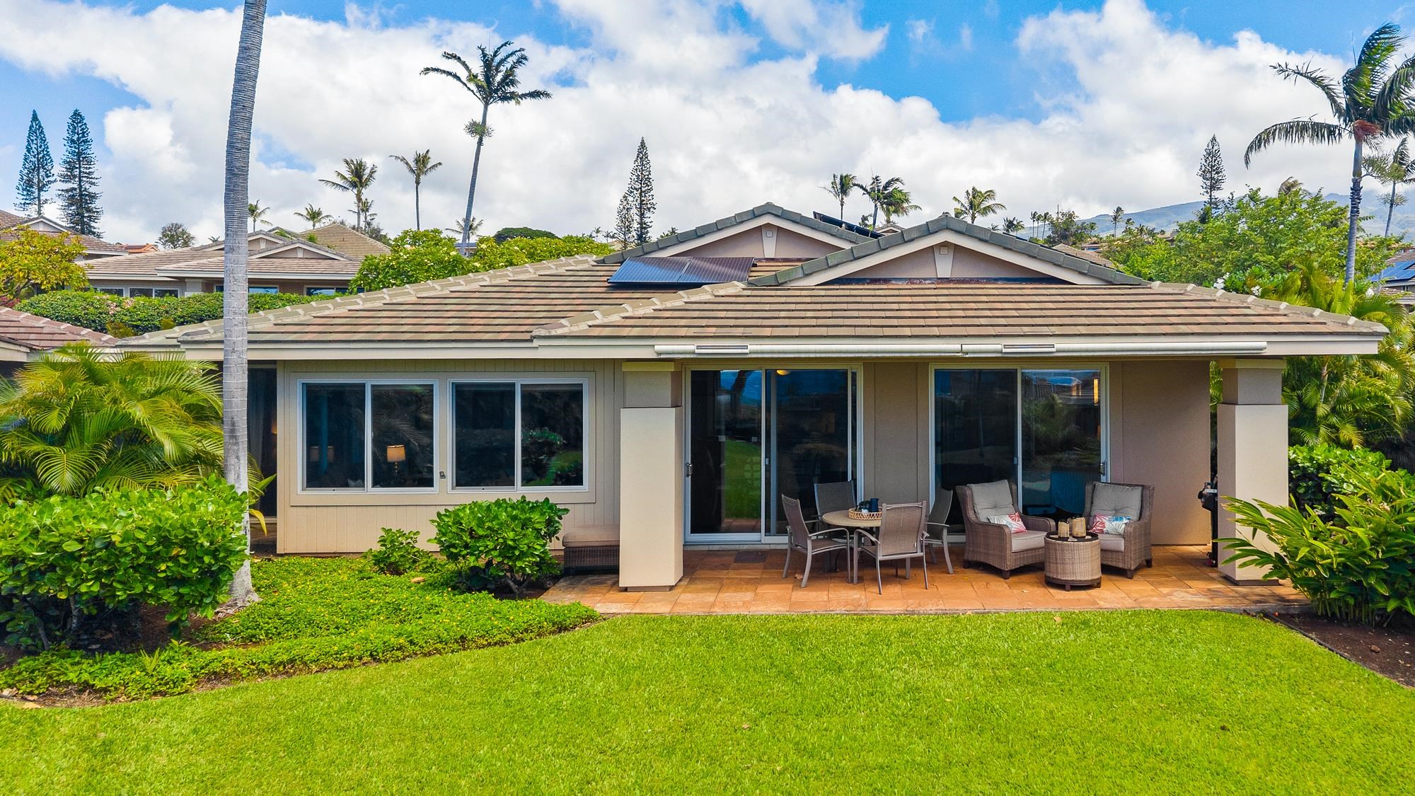 Home exterior with solar panels, a furnished covered patio, tropical landscaping, and distant hills.