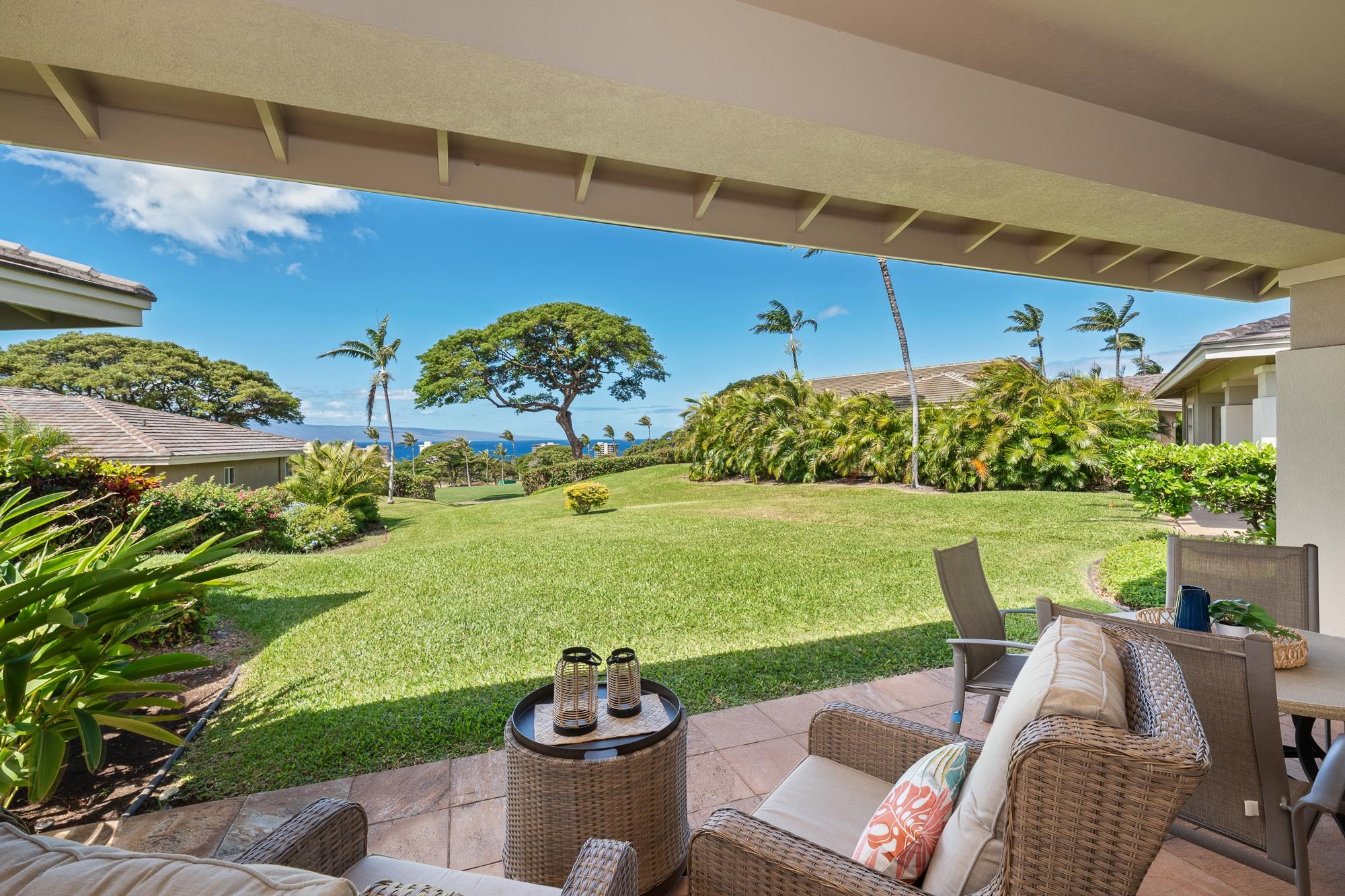 Covered patio with wicker seating overlooks a green lawn, palm trees, and the ocean.