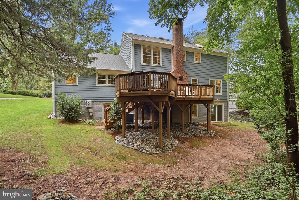 Rear of a gray two-story house with a raised wooden deck and brick chimney, surrounded by trees.