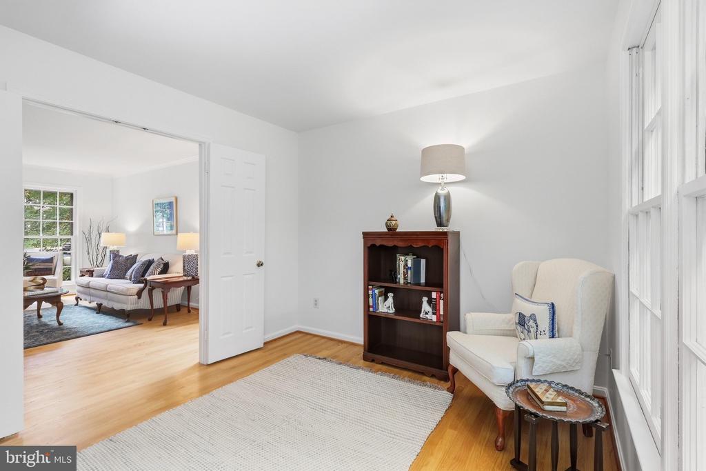 Furnished study with a wooden bookcase and armchair beside tall windows, opening onto the living room.