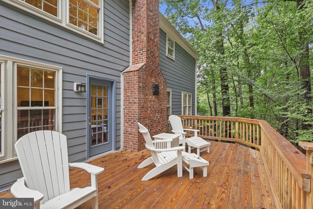 Wooden deck with white Adirondack chairs beside a gray-sided home, overlooking leafy woods.
