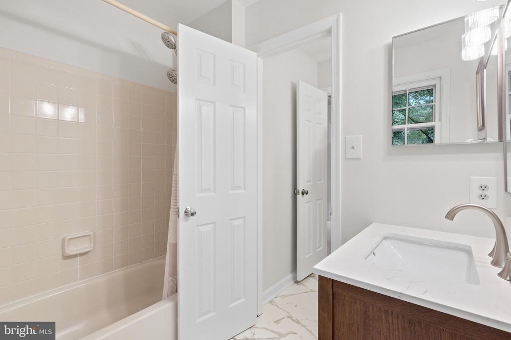 Bathroom with beige-tiled tub and shower, white vanity, and window overlooking greenery.
