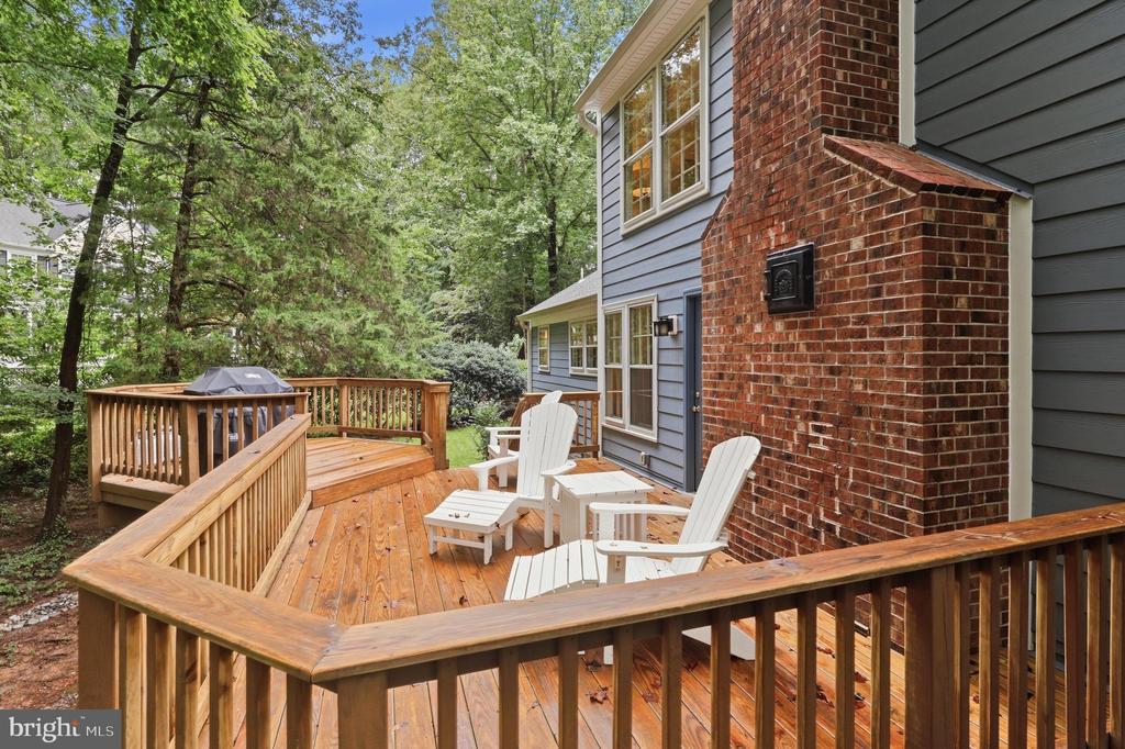 Wooden rear deck with white Adirondack chairs beside a brick chimney, surrounded by mature trees.