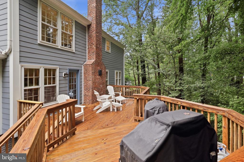 Wooden backyard deck with Adirondack chairs and covered grills beside a gray-sided home and brick chimney.