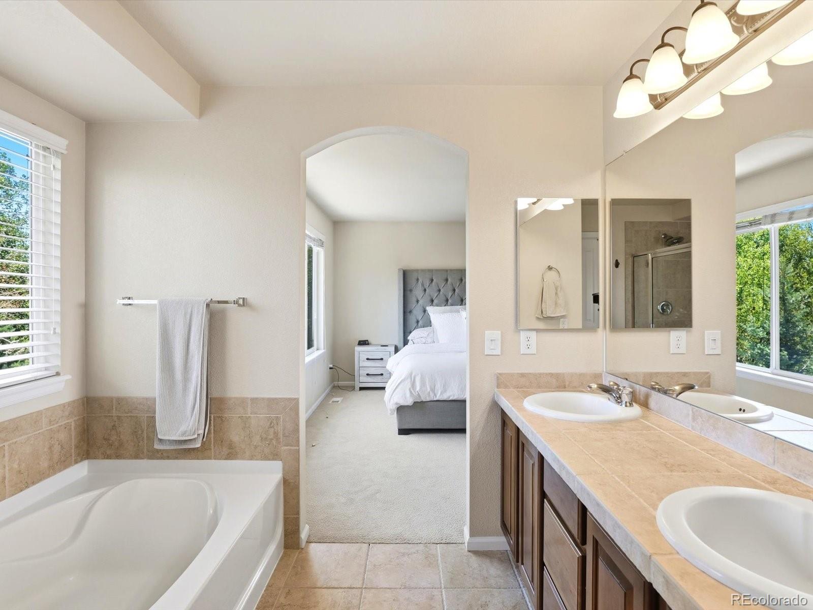Primary bathroom with soaking tub and double vanity, opening to a bedroom with a gray upholstered headboard.