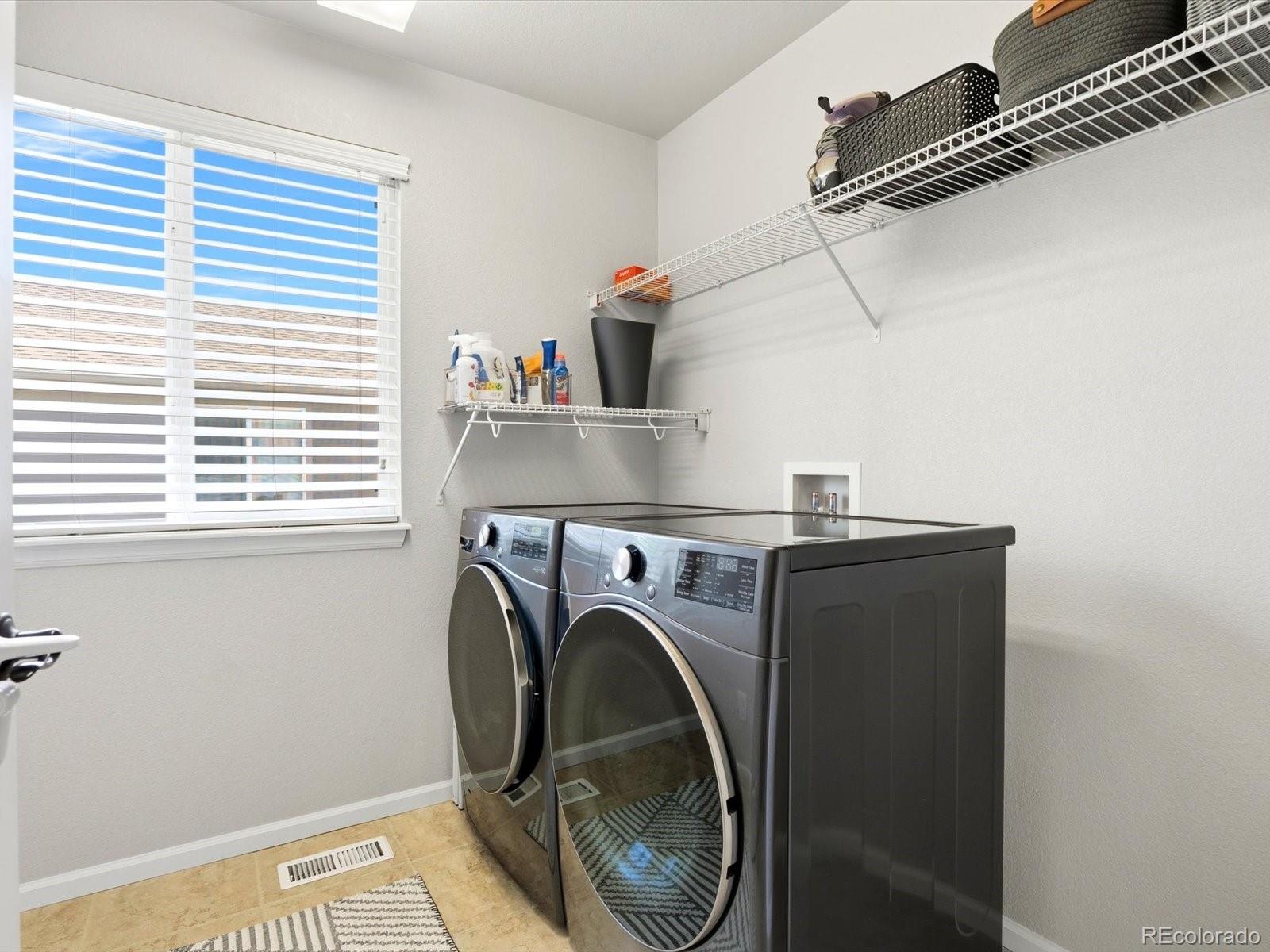 Laundry room with front-loading washer and dryer beneath wire shelves, beside a window with blinds.