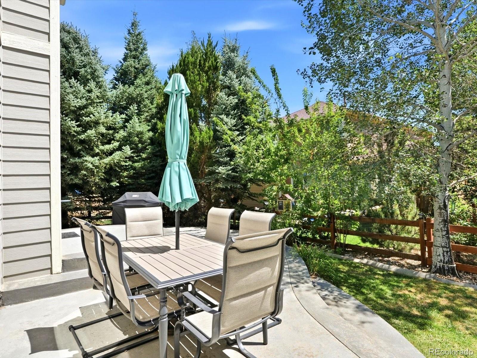 Backyard patio with outdoor dining set and turquoise umbrella, bordered by mature trees and landscaping.