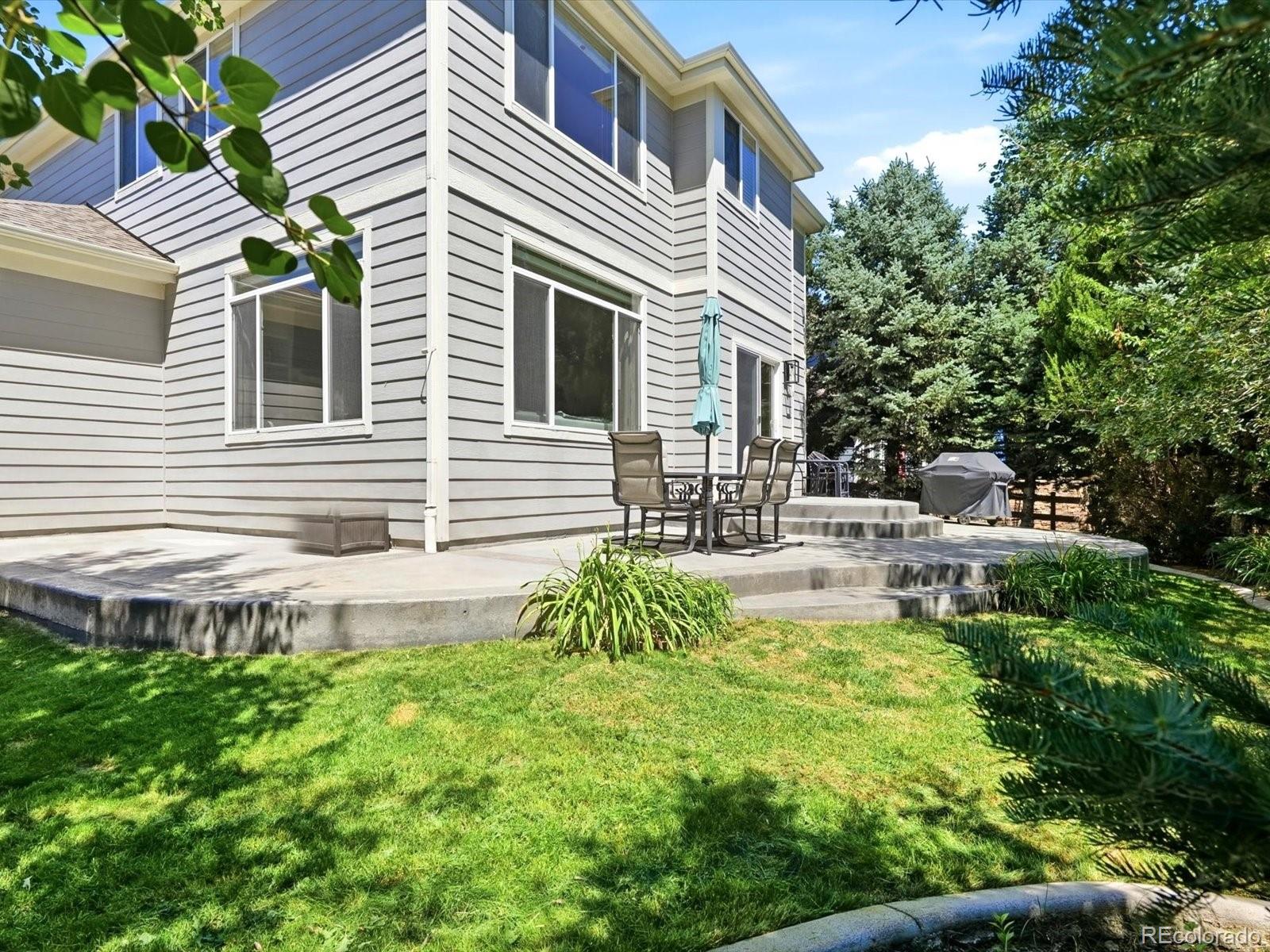 Gray two-story home with a raised patio, outdoor chairs, closed umbrella, and leafy lawn.