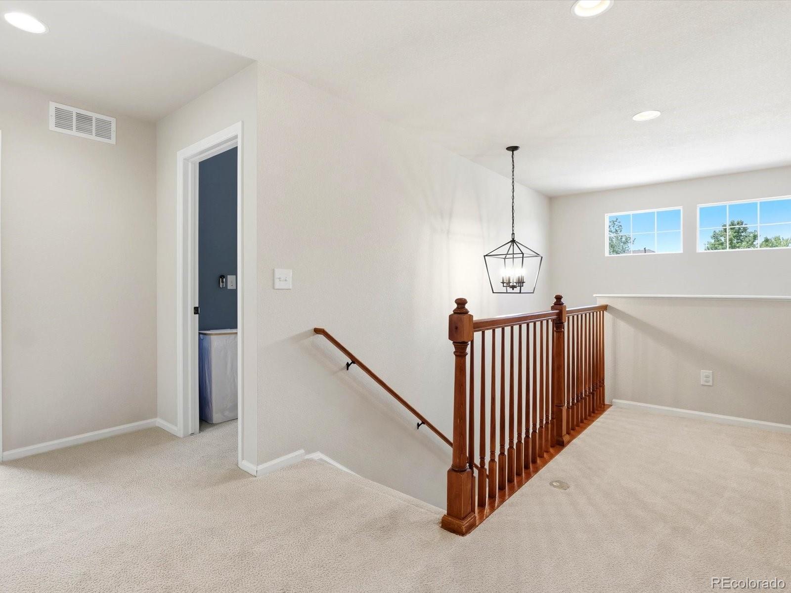 Upstairs landing with beige carpet, wood stair railing, black chandelier, and two high windows.