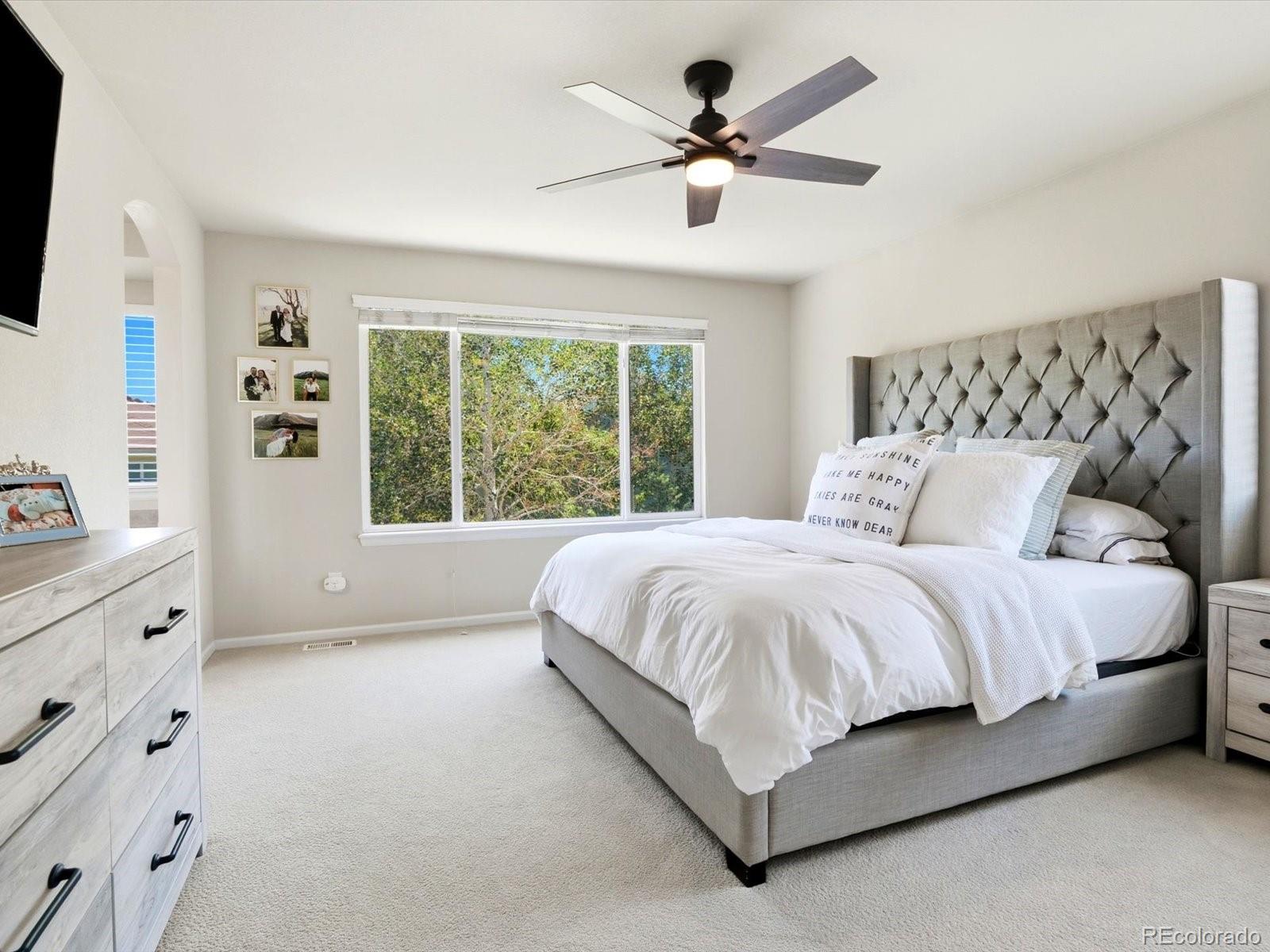 Bedroom with a gray tufted bed, white bedding, and a large window overlooking trees.
