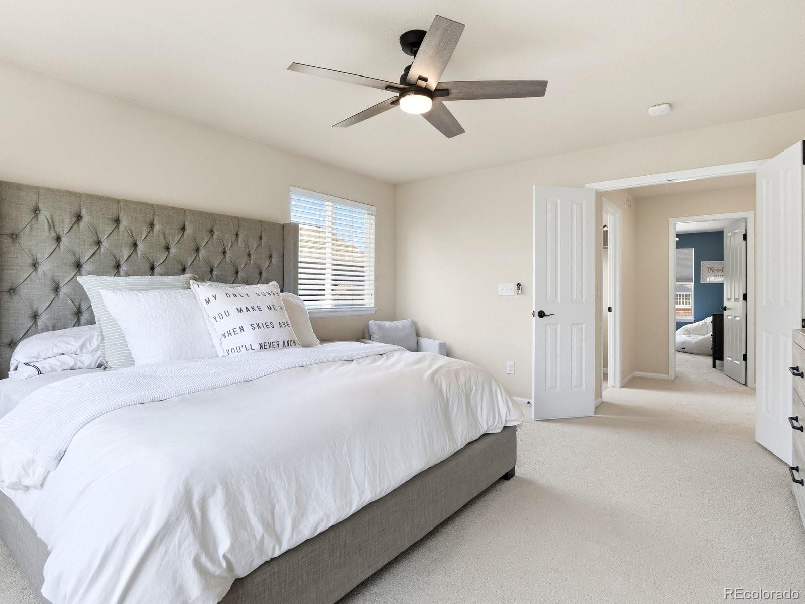Bedroom with a white bed, gray tufted headboard, ceiling fan, and open doors to a hallway.