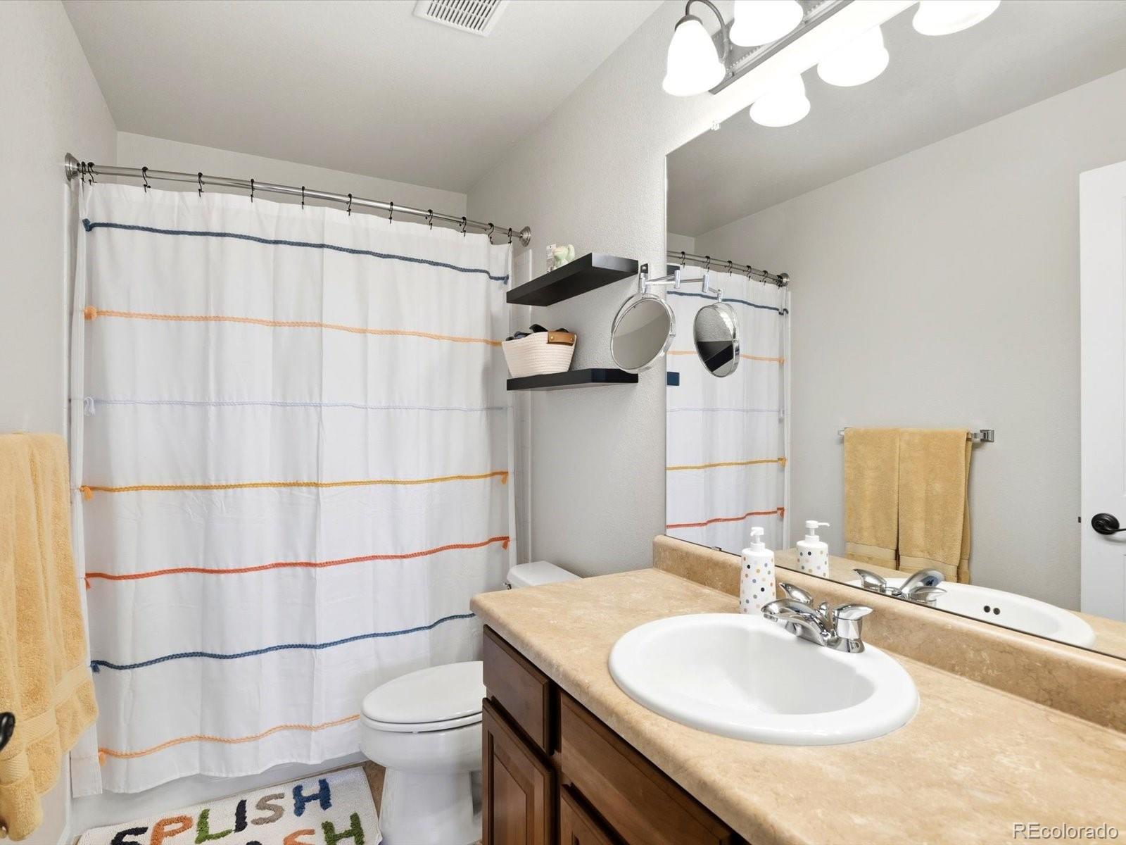 Bathroom with a striped shower curtain, toilet, and double-sink vanity with beige countertops.