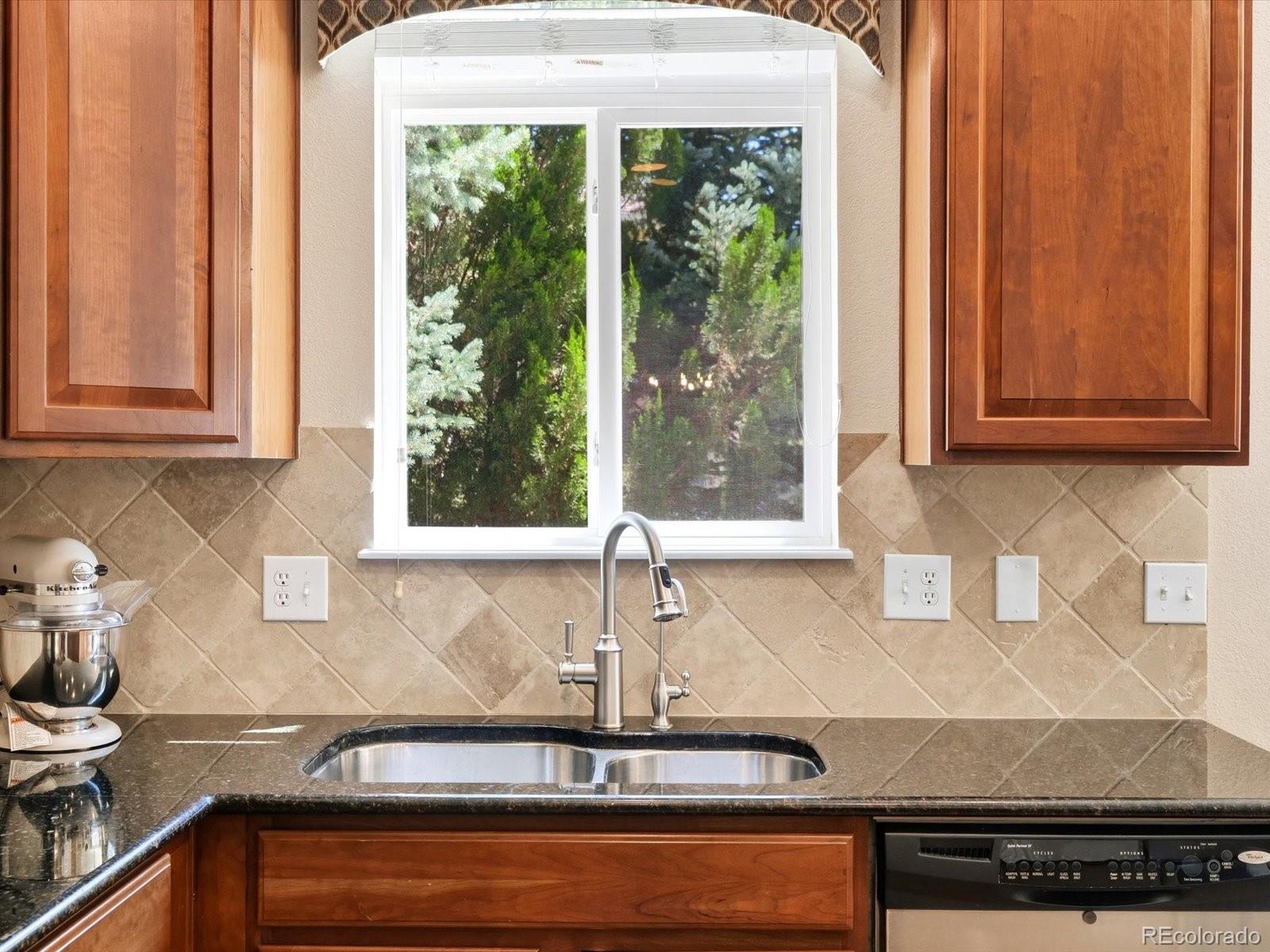 Kitchen sink beneath a window, framed by wood cabinets and a tan diamond-pattern tile backsplash.