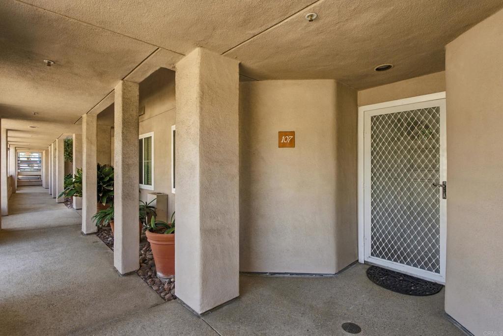 Condominium entry 107 with a white security screen door and a covered corridor lined with potted plants.